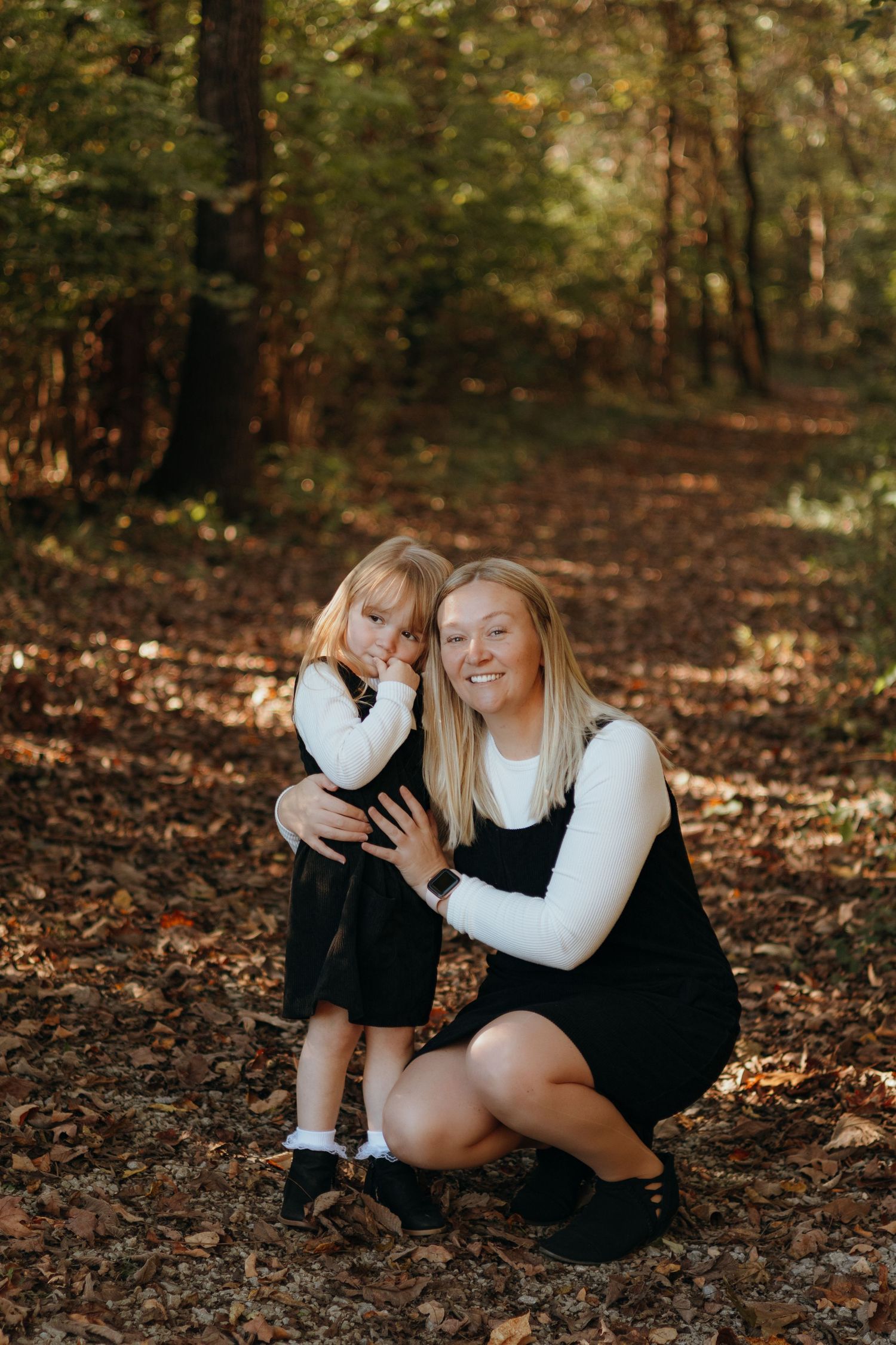 Smiling mother in a black dress crouches in an autumn forest hugging her little daughter among fallen leaves.