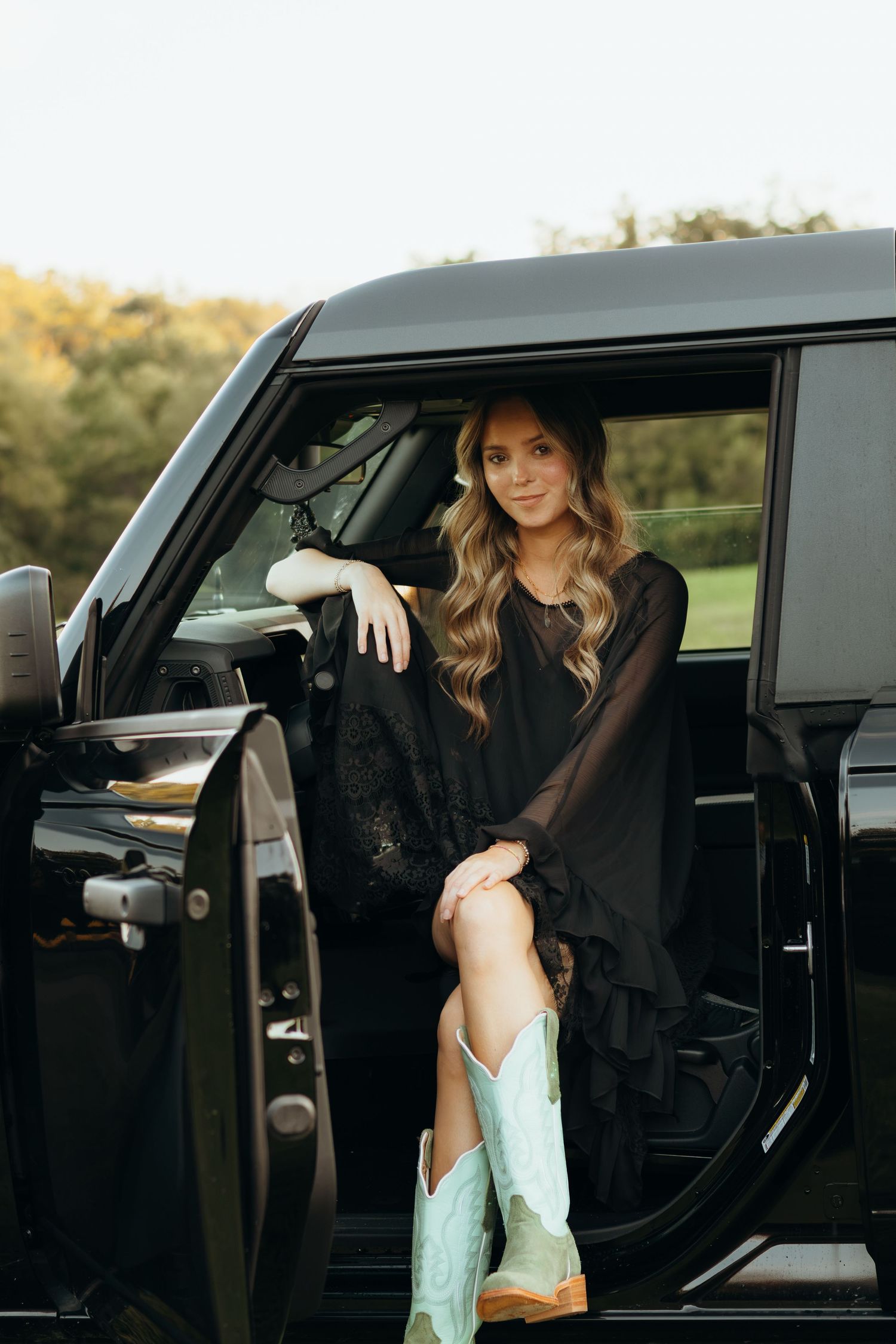 Woman with long wavy hair in a black ruffled dress and turquoise cowboy boots sitting in the open door of a black SUV outdoors.