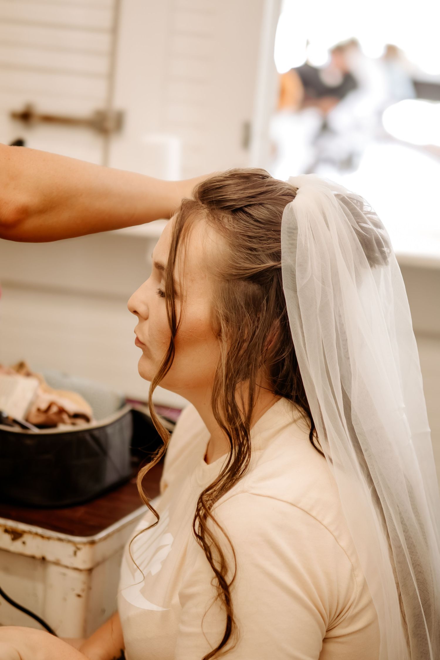 A bride with loose wavy hair sits with eyes closed while a stylist pins her long white veil during wedding morning preparations.