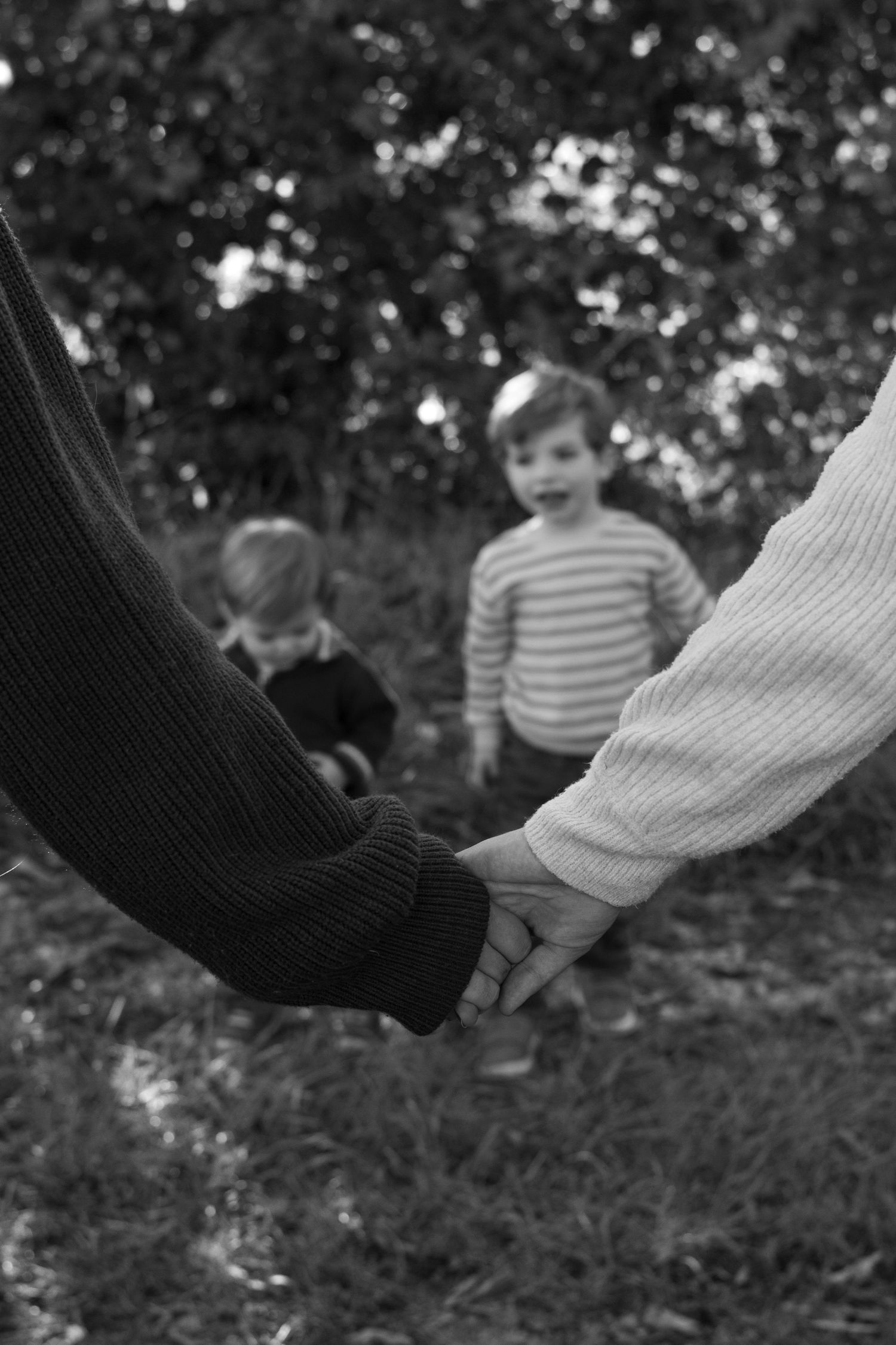 Black and white photo of two adults holding hands in the foreground while two young children stand on the grass among trees.