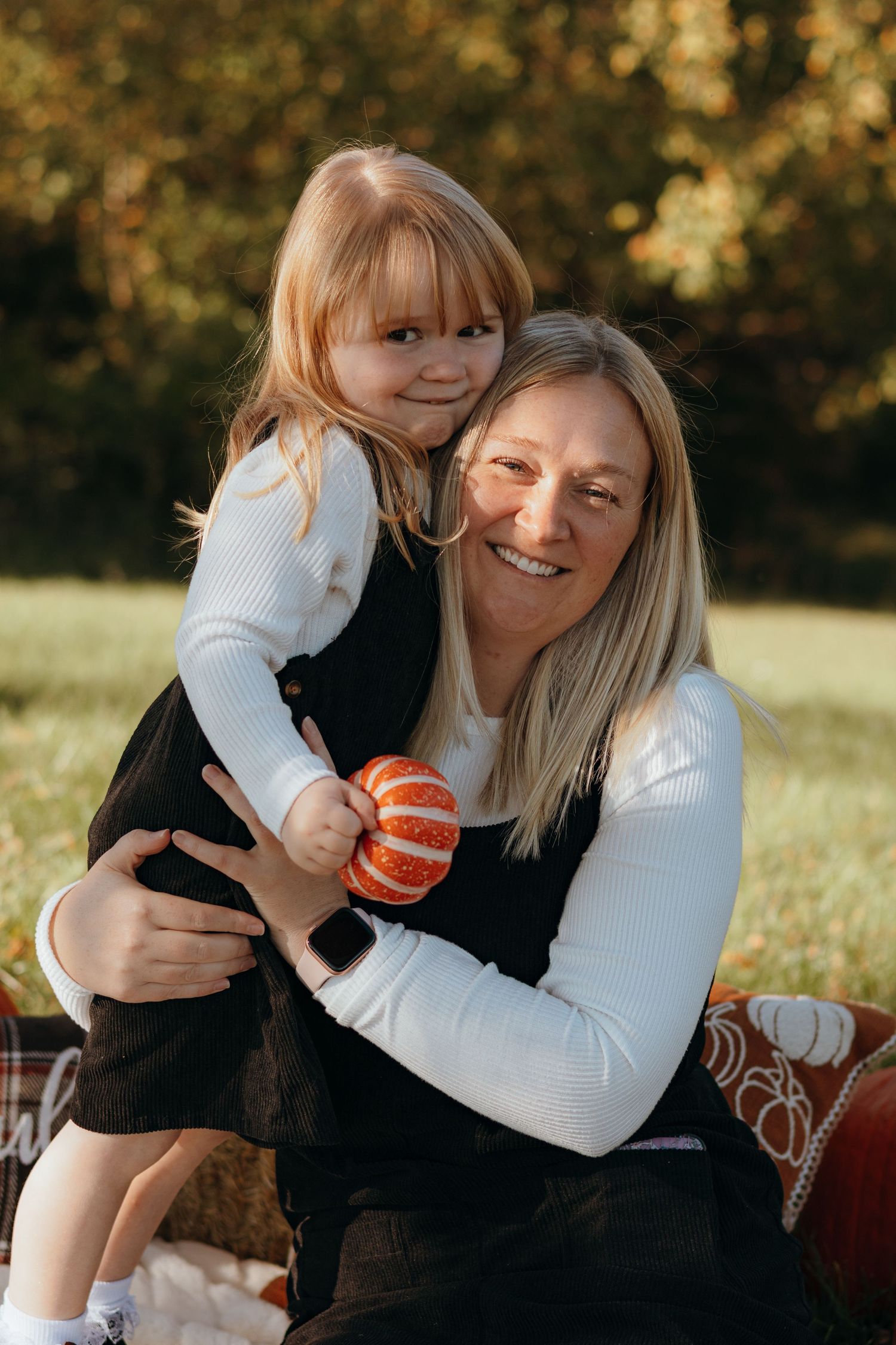 Smiling woman with blonde hair hugs a young girl holding a small orange pumpkin on a blanket in an autumn park.