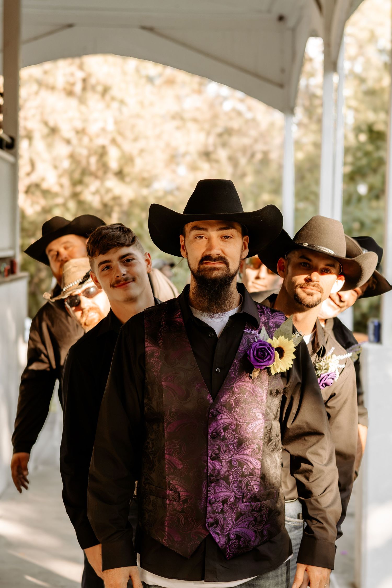Groomsmen in black cowboy hats and purple paisley vests with floral boutonnieres line up outside at a rustic wedding venue.