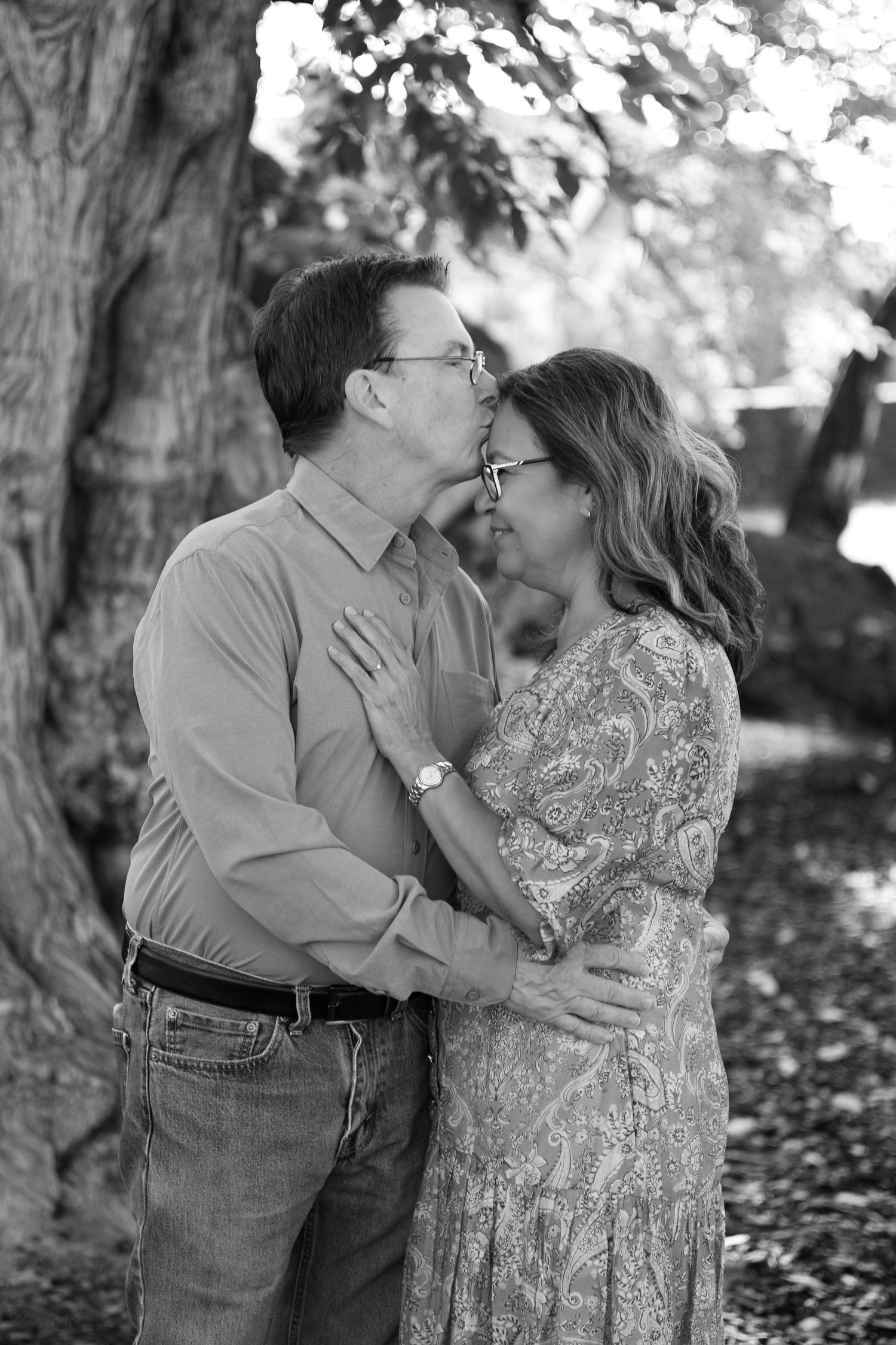 Black and white photo of a couple embracing outdoors as a man kisses the forehead of a smiling woman beside a large tree.