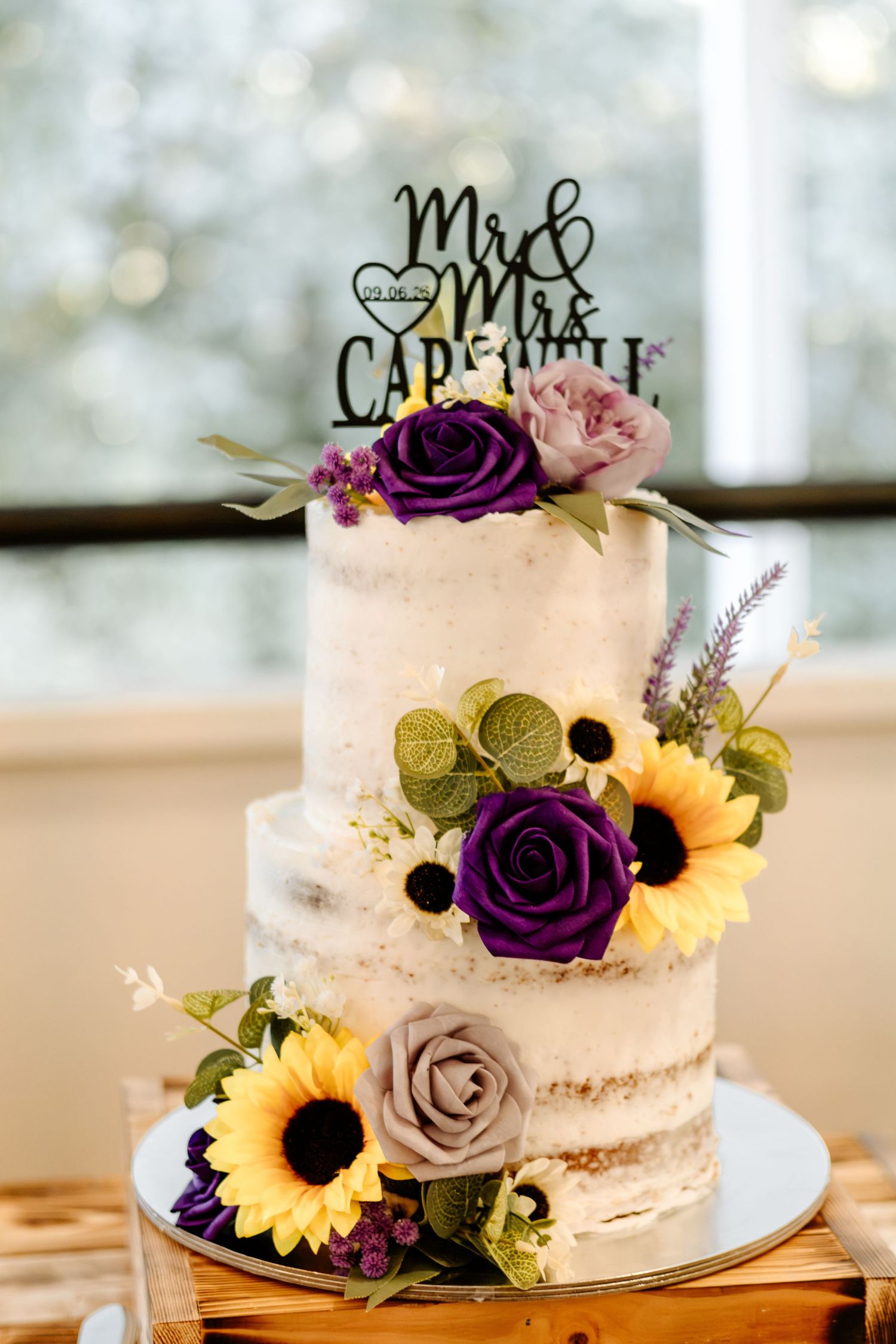 Two-tier rustic naked wedding cake decorated with sunflowers, purple roses and greenery, topped with a Mr and Mrs cake topper.