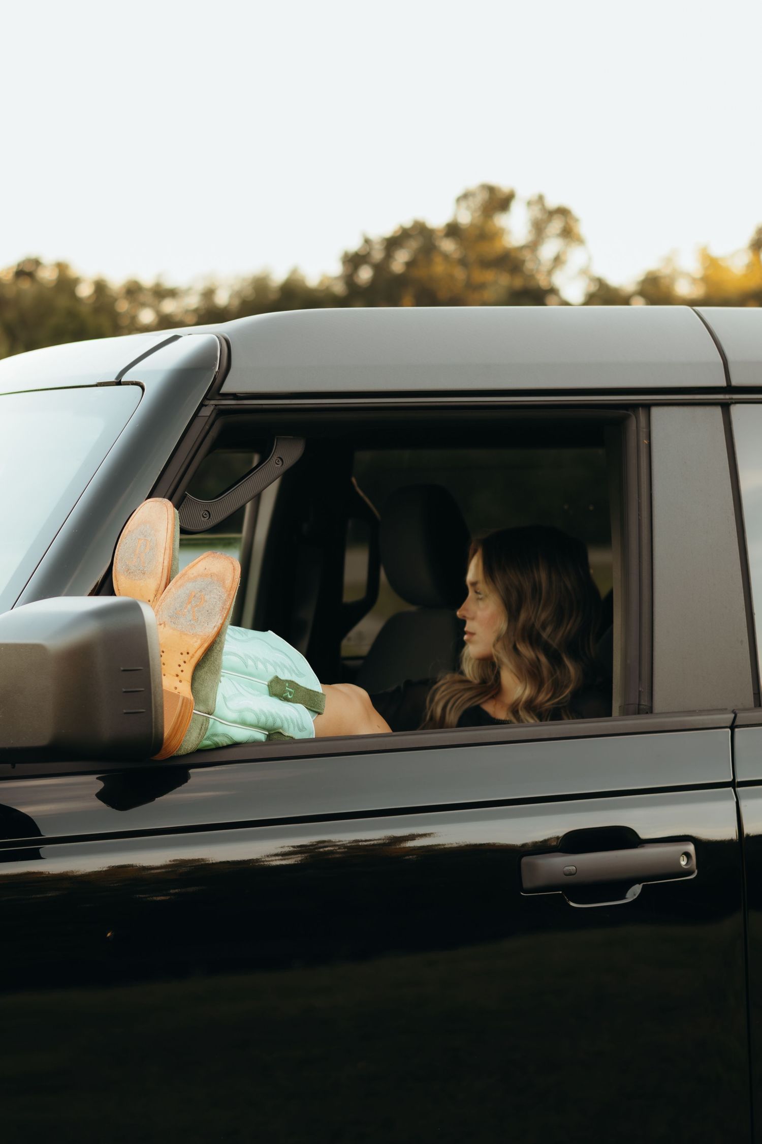 Woman with wavy hair sits in a black SUV with her mint green cowboy boots propped out the open passenger window at sunset.