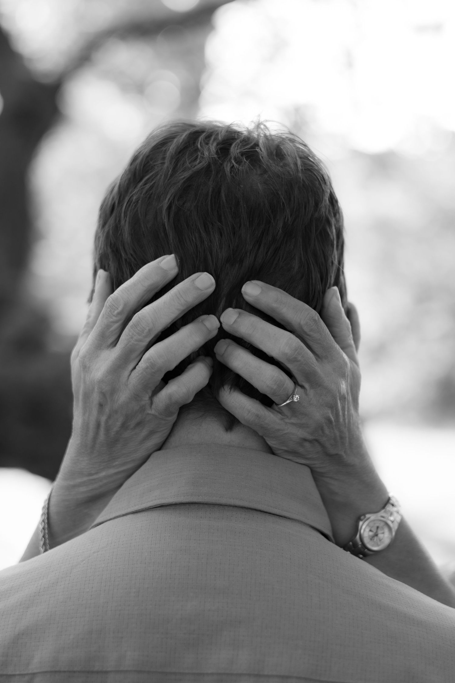 Black and white photo from behind of a man with a woman's hands gently cradling his head, wearing a ring and watch.
