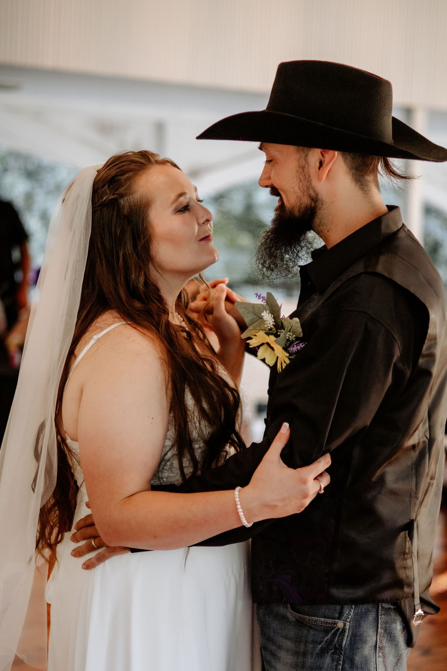 Bride in a white gown and veil shares a close first dance with a bearded groom in a black cowboy hat and western-style wedding attire.