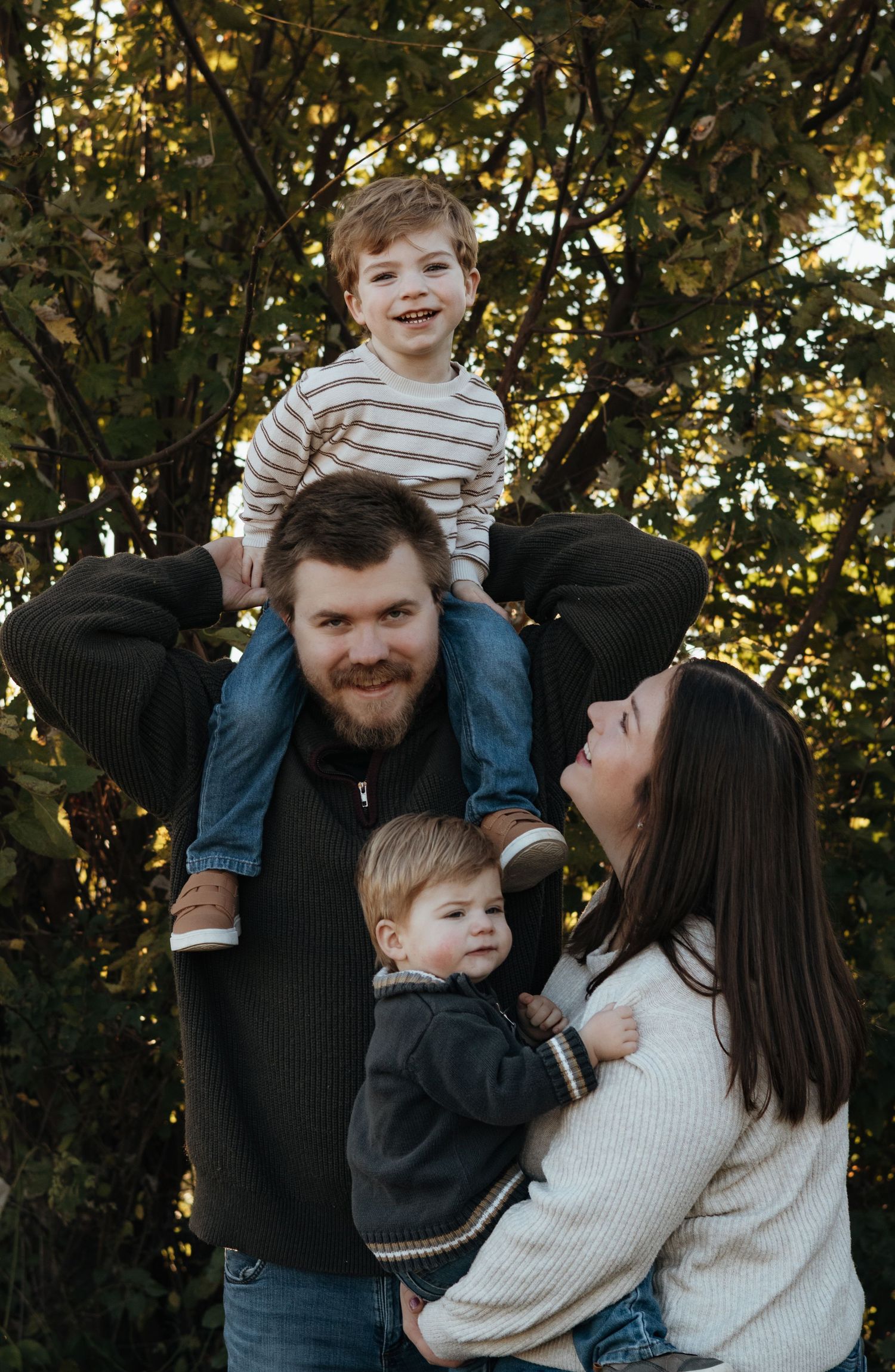 Smiling family portrait outdoors with a bearded man carrying a toddler on his shoulders while a woman holds a baby among autumn trees.