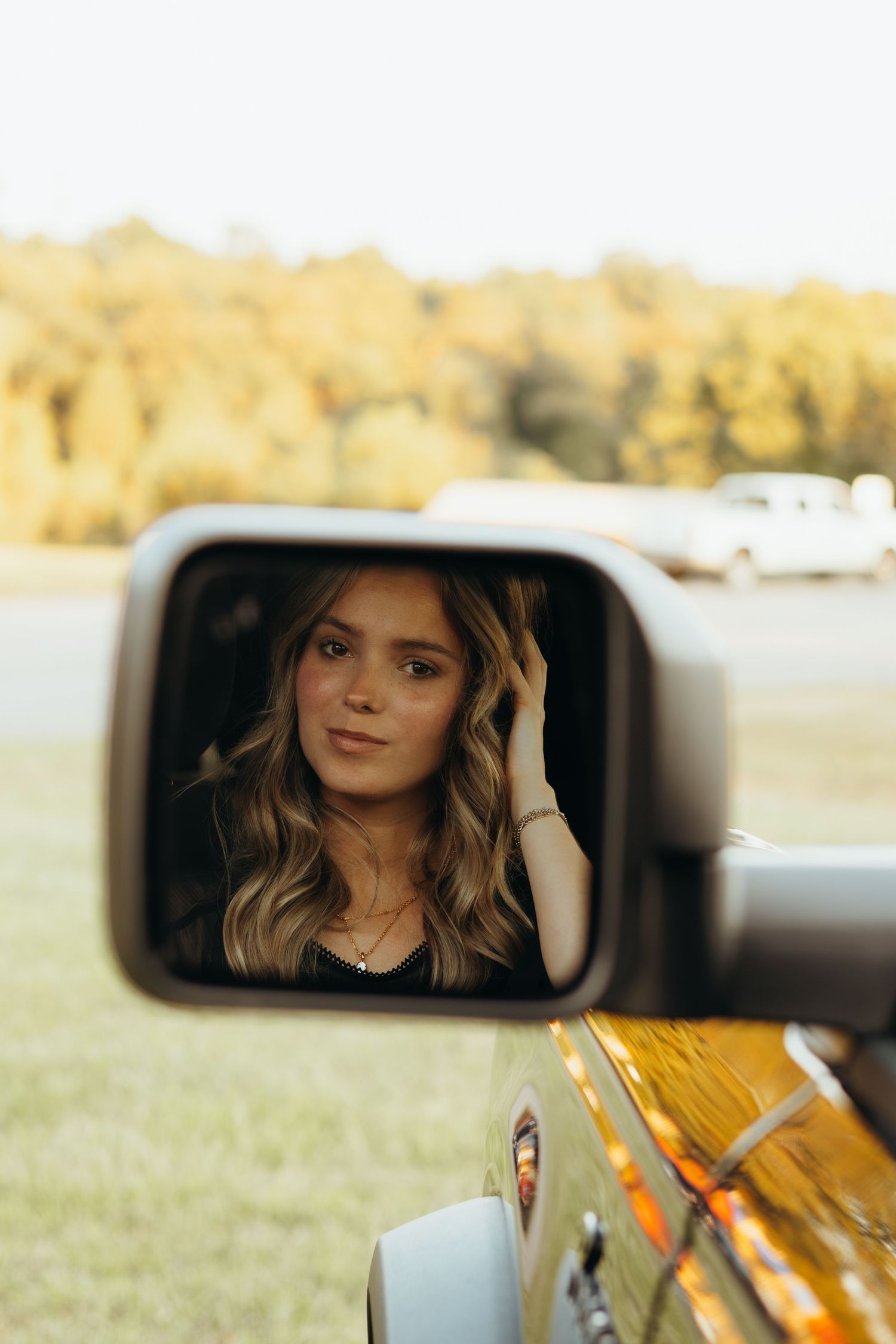 Young woman with wavy hair reflected in a car side mirror, touching her hair, with golden autumn trees and a grassy field behind.