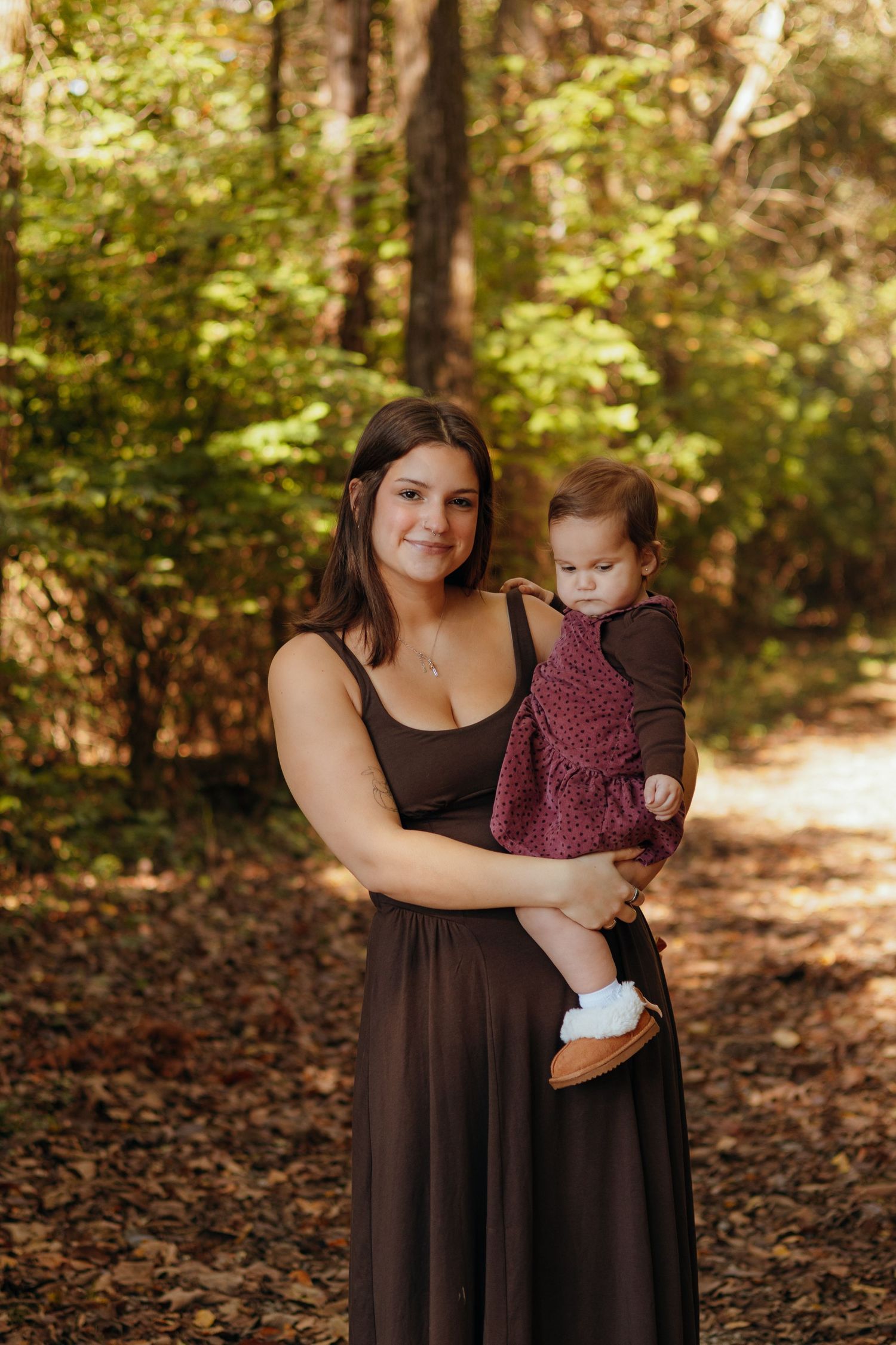 Smiling woman in a brown dress holds a toddler in a burgundy dress on a sunlit forest path covered in fallen autumn leaves.