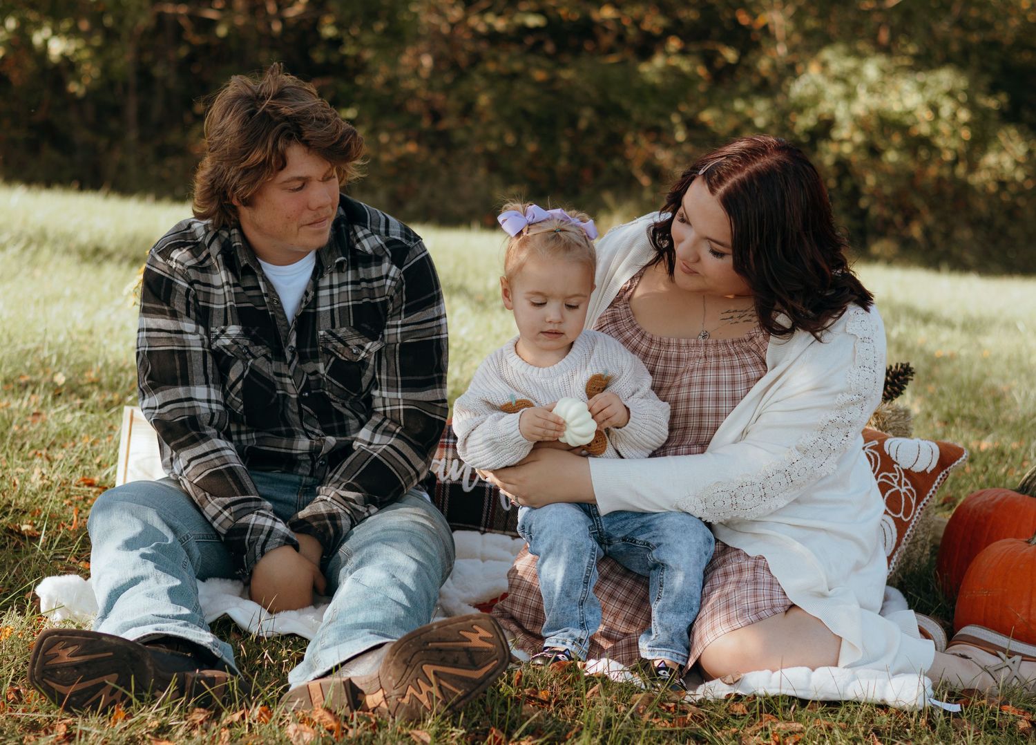 Family of three relaxes on a picnic blanket in a grassy field with pumpkins, parents smiling as they hold their baby girl.