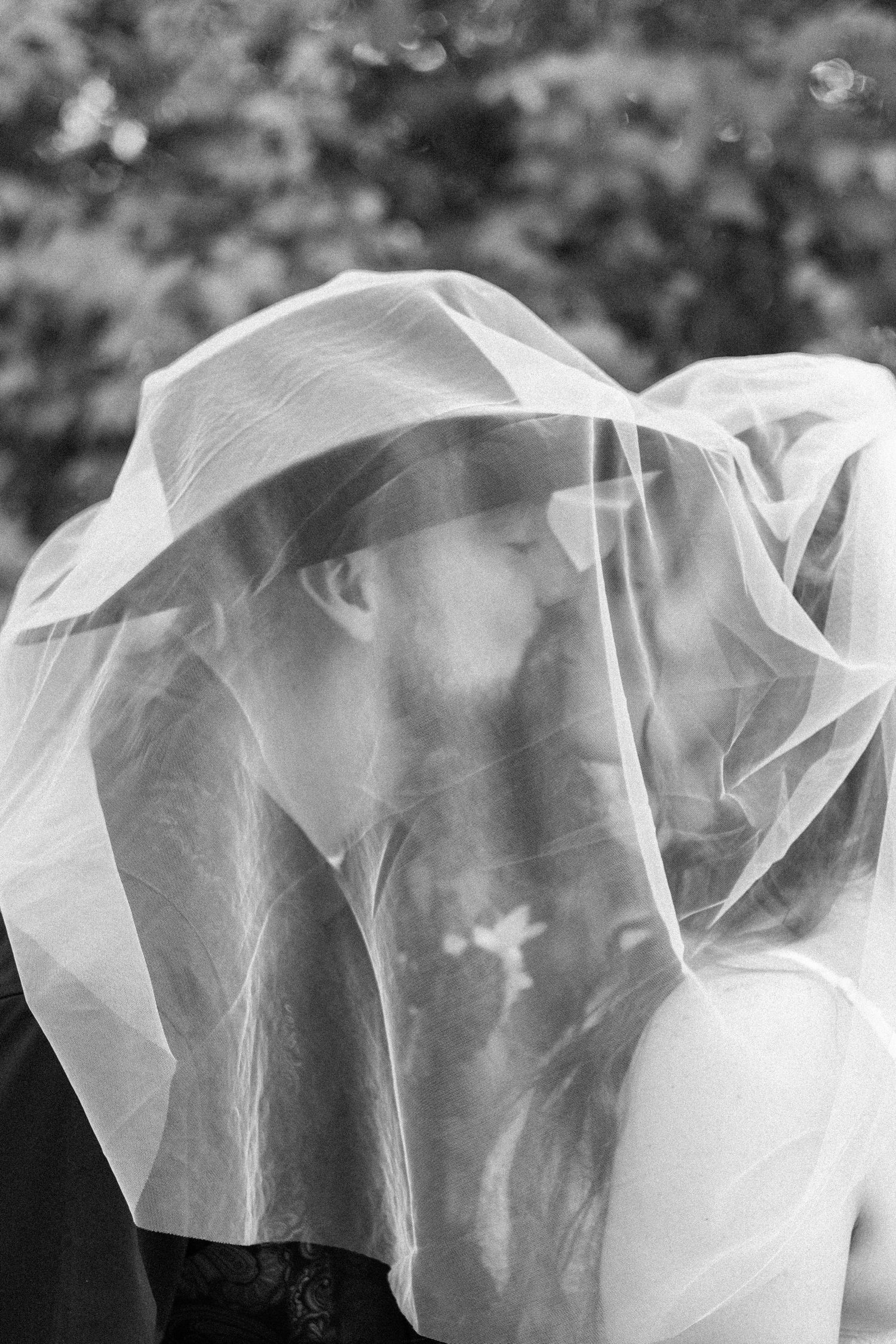 Black and white wedding photo of a couple kissing beneath a sheer bridal veil, with soft blurred greenery in the background.
