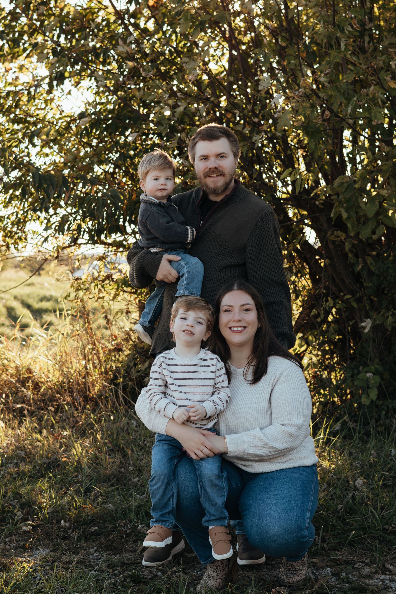 A smiling family of four poses outdoors in autumn, with a bearded father holding one toddler and a mother kneeling beside another child.