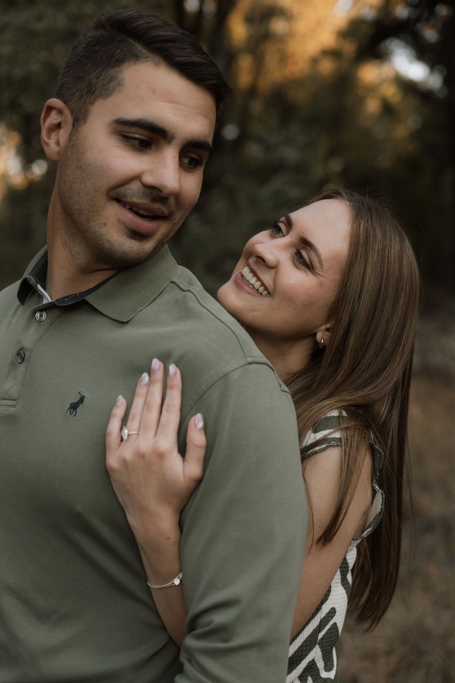 A woman with long brown hair smiles warmly as she hugs a man from behind, wearing an engagement ring, in soft golden forest light.