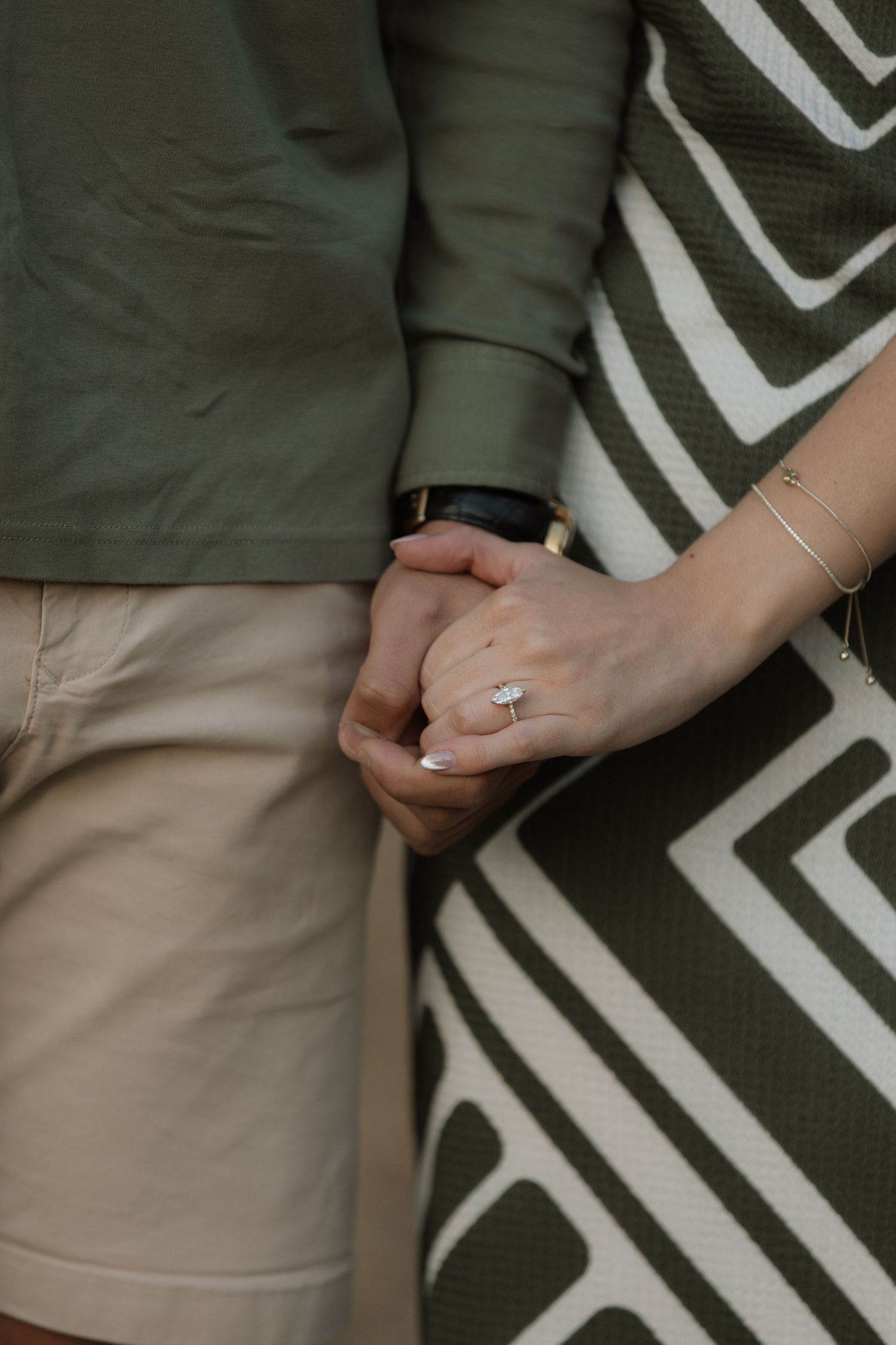 Close-up of a couple holding hands, the woman's sparkling engagement ring and gold bracelets visible against a green patterned dress.