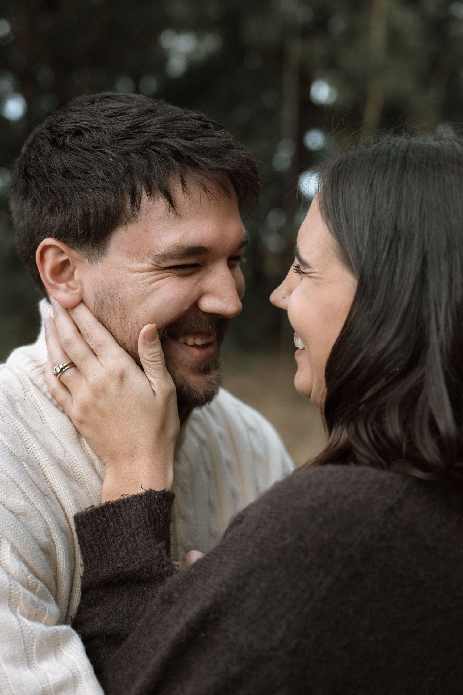 Smiling husband and wife share a tender moment as she gently touches his face, with a blurred forest background behind them.