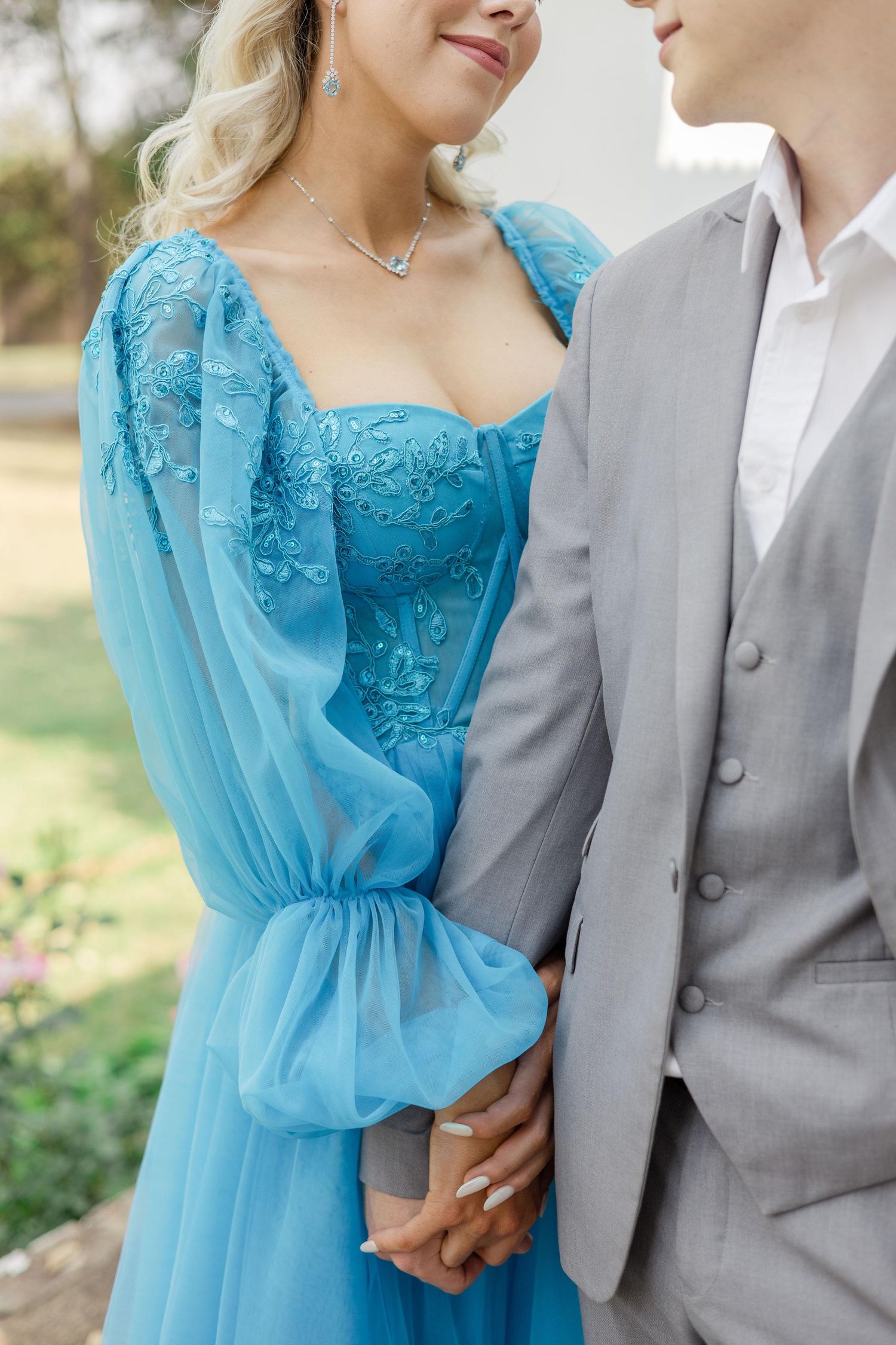 Couple holding hands outdoors, the woman in a blue embroidered dress and the man in a light gray suit vest and white shirt.
