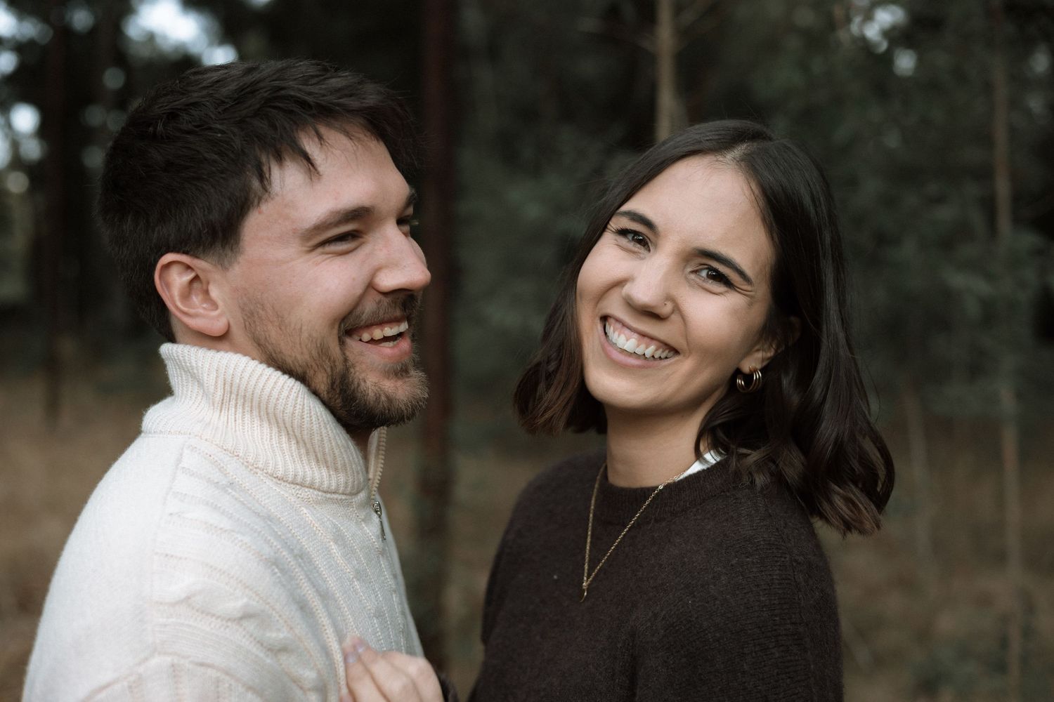 Happy couple laughing together outdoors in cozy sweaters during a relaxed engagement photoshoot among the trees.