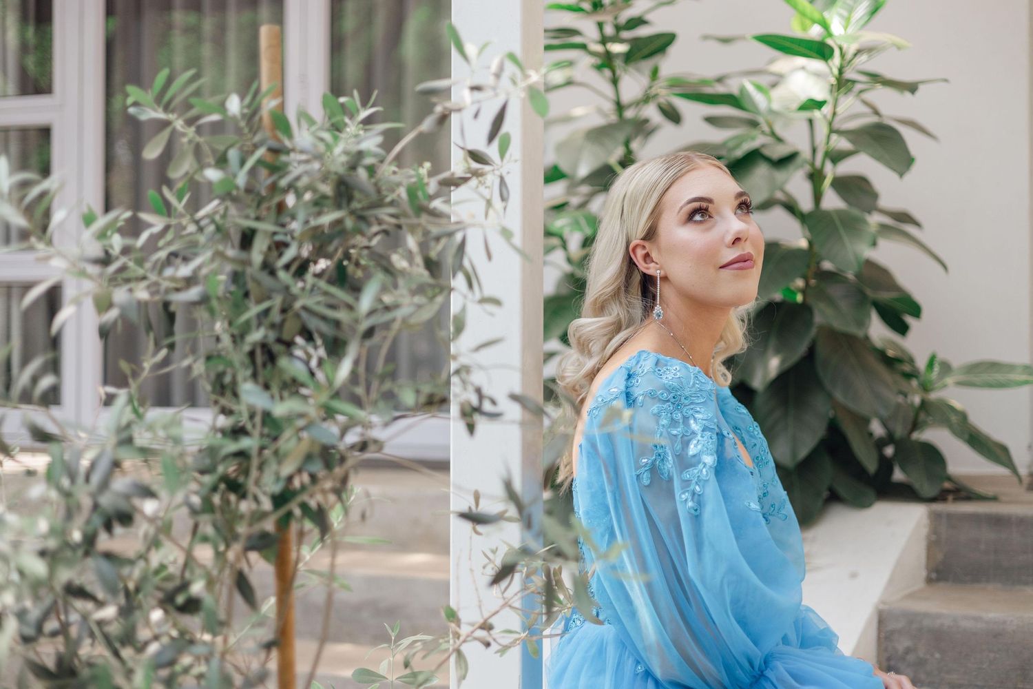 Woman in a flowing blue embroidered dress sits beside lush green olive trees in a bright, romantic garden portrait.