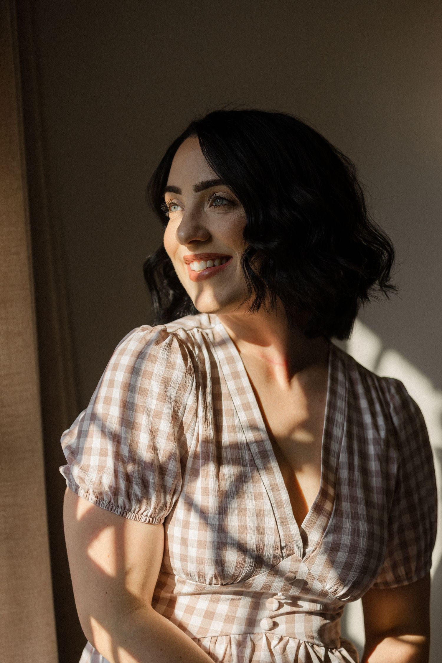 Smiling woman with short dark wavy hair wears a pink gingham puff-sleeve blouse, lit by warm golden sunlight streaming through a window.