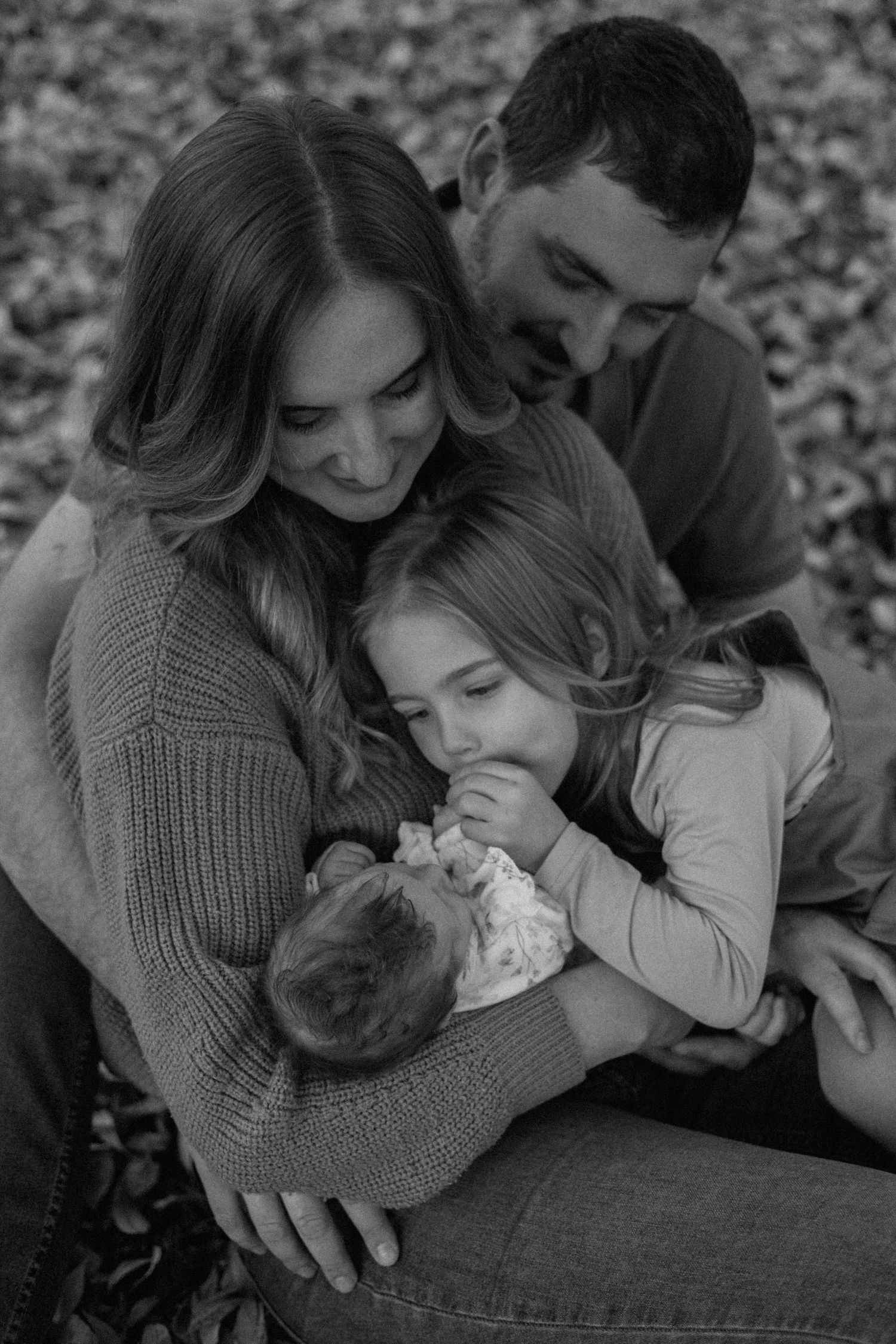 Black and white photo of a mother, father and young daughter cuddling a sleeping newborn baby on a bed of autumn leaves.