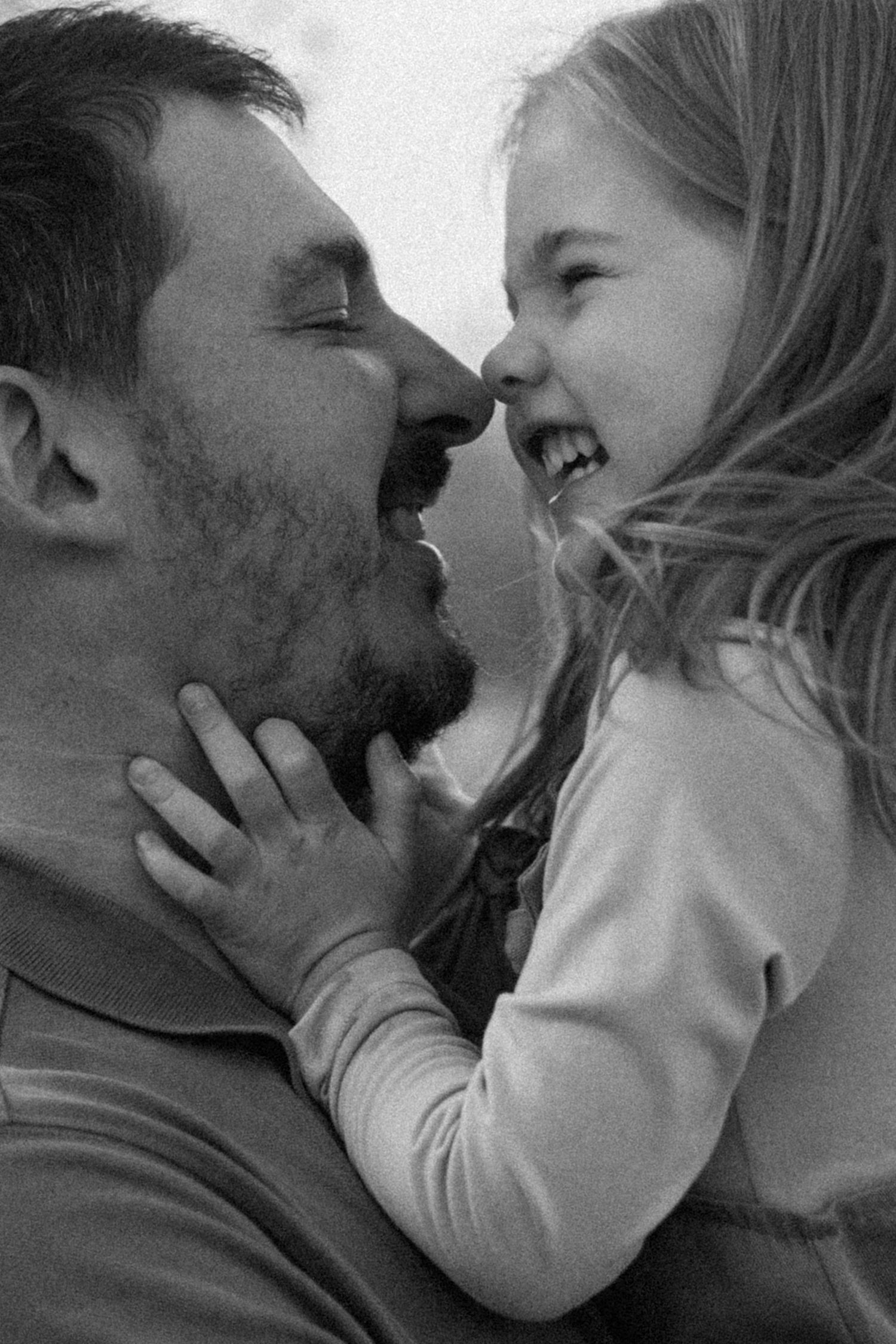 Black and white close-up of a bearded father and his laughing young daughter touching noses in an affectionate moment.