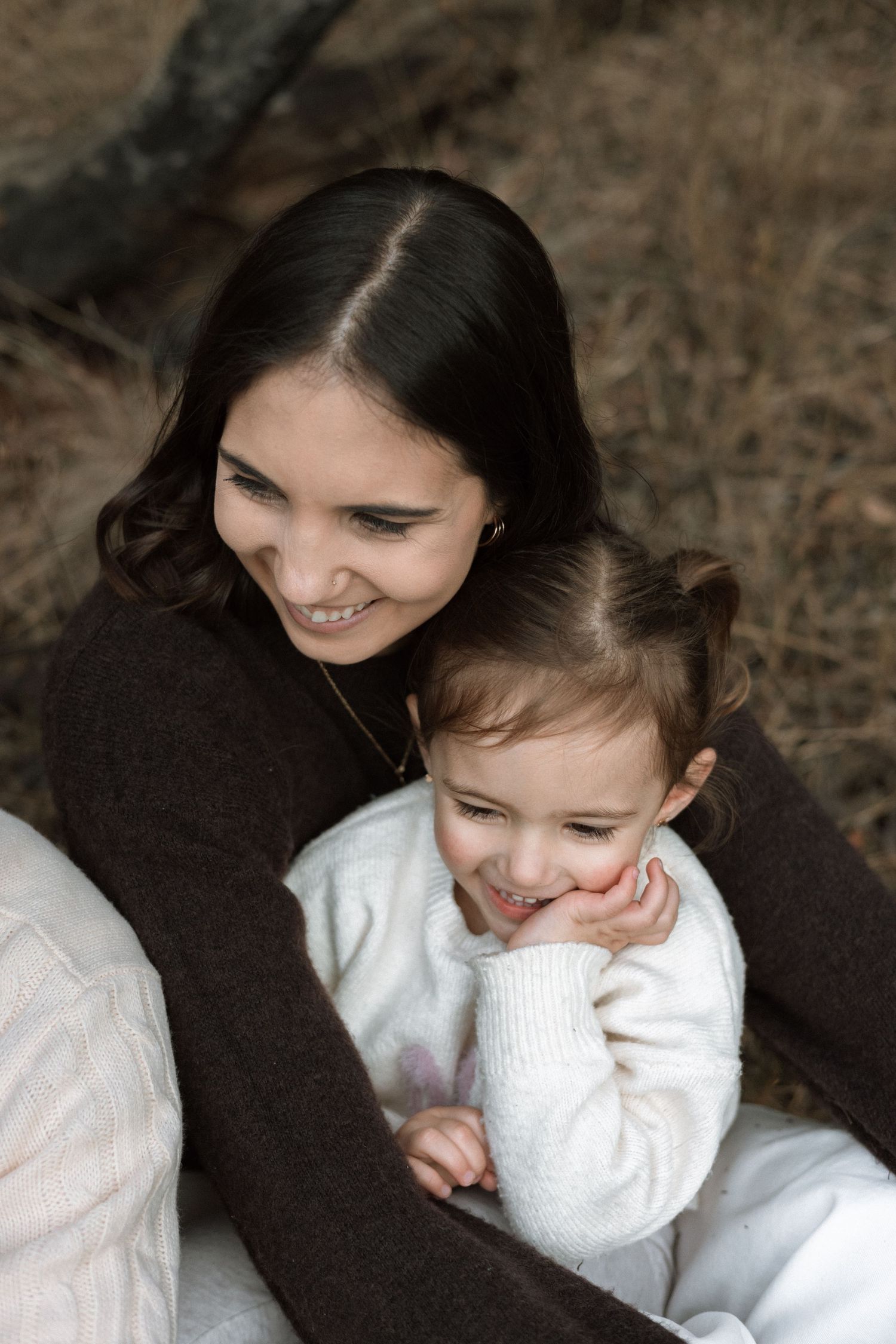 Dark-haired mother laughs while hugging her giggling toddler daughter on her lap outdoors during a family photo session.