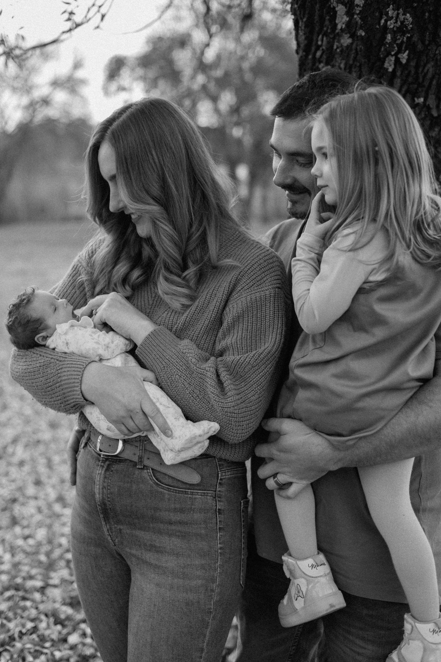 Black and white family portrait of parents with a newborn baby and a young daughter in her father's arms beside a large tree.
