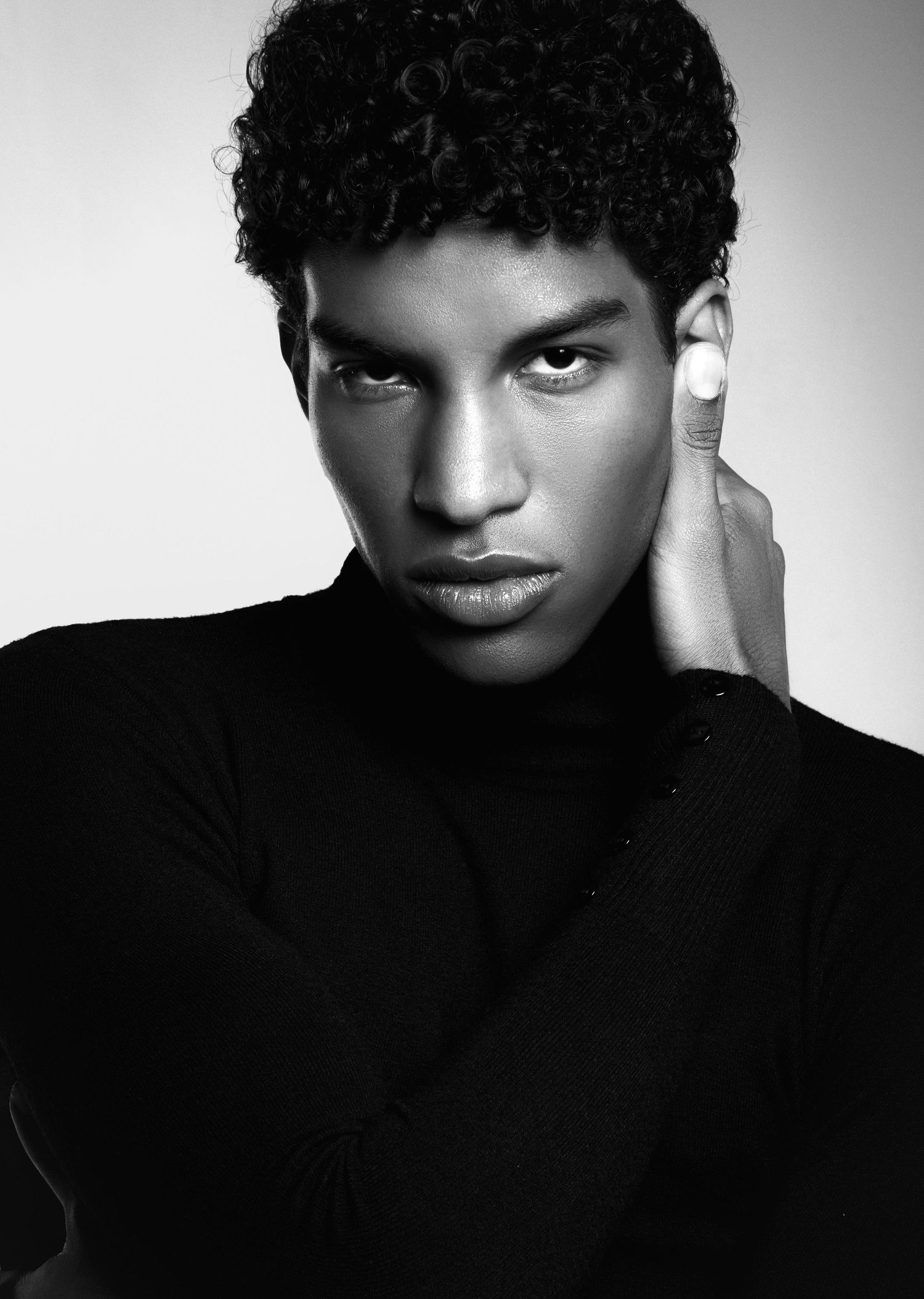 Black and white portrait of a young man with curly hair in a black turtleneck, touching his neck with an intense gaze.
