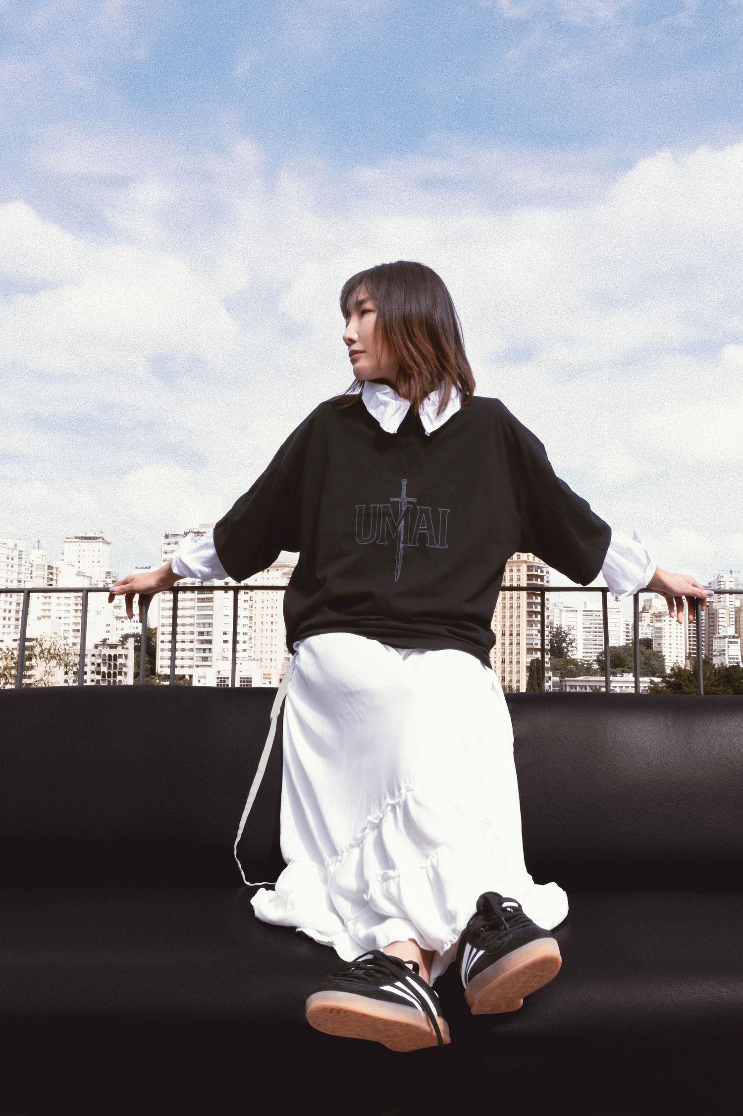 Woman in profile wears a black graphic sweatshirt, white ruffled skirt and sneakers while seated on a rooftop couch under a cloudy sky.