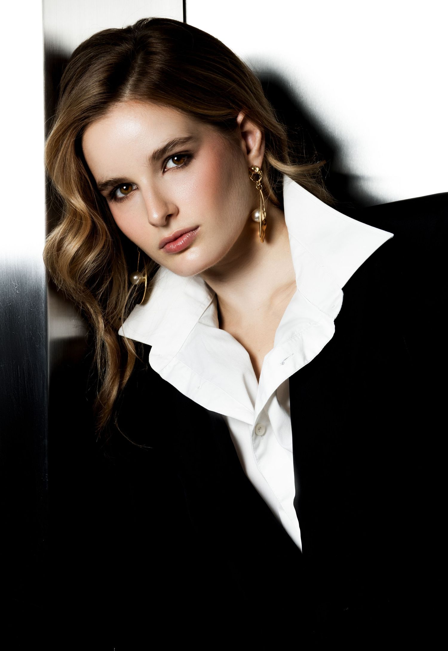 Portrait of a model with wavy brown hair wearing gold drop earrings, a white collared shirt and a black blazer in dramatic light.