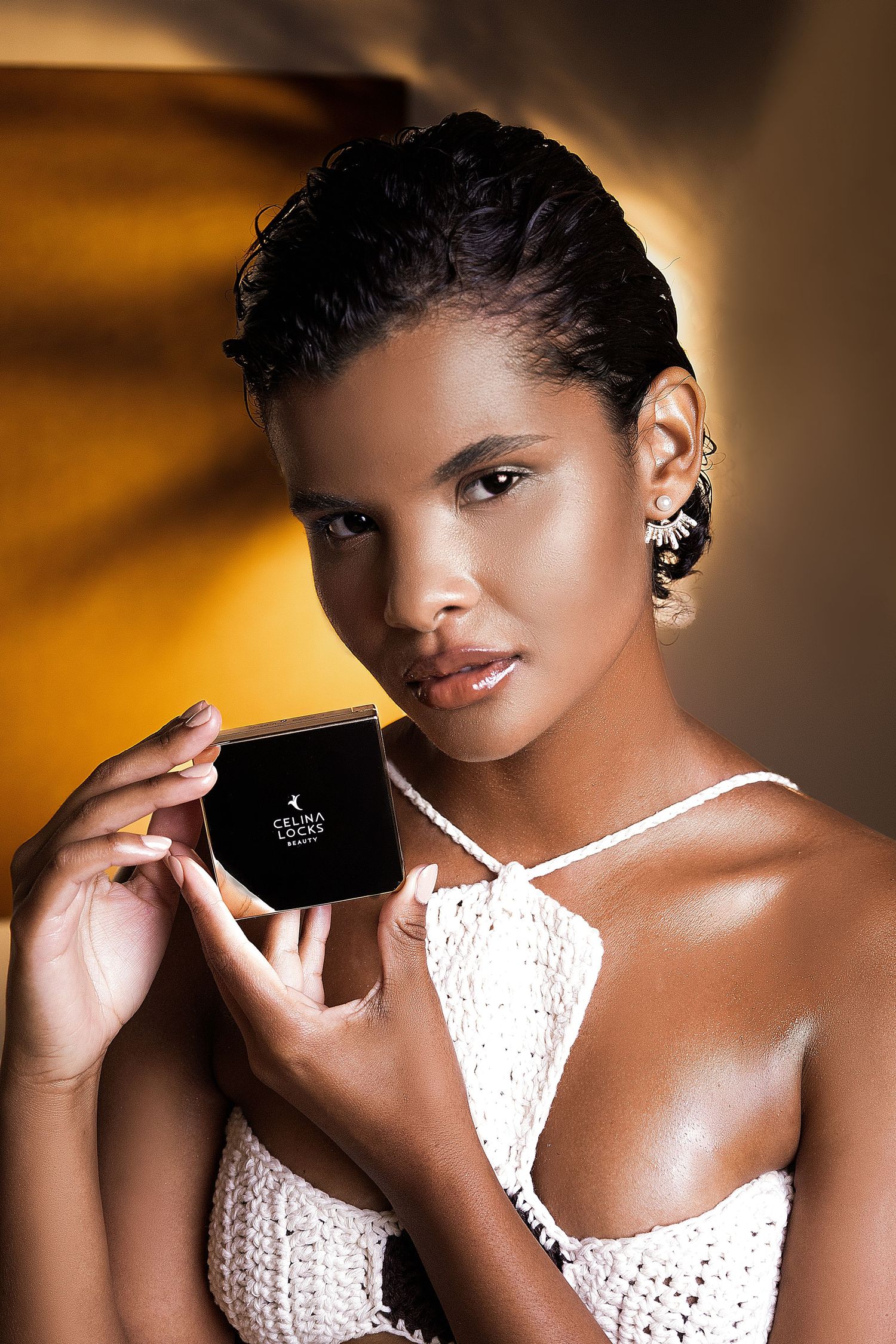 Woman with short hair and glowing skin in a white crochet halter top holds a black luxury beauty product box in warm golden light.
