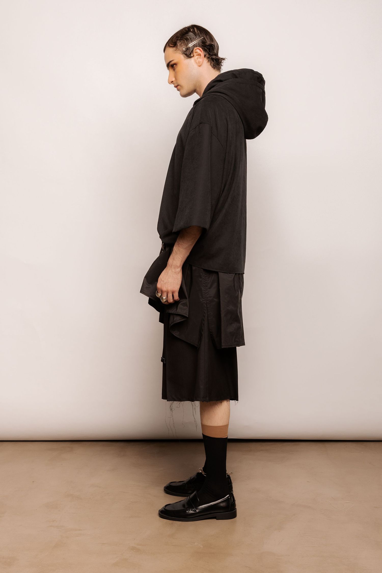 Side view of a black oversized hooded top with layered asymmetric black shorts, black socks and glossy black loafers.