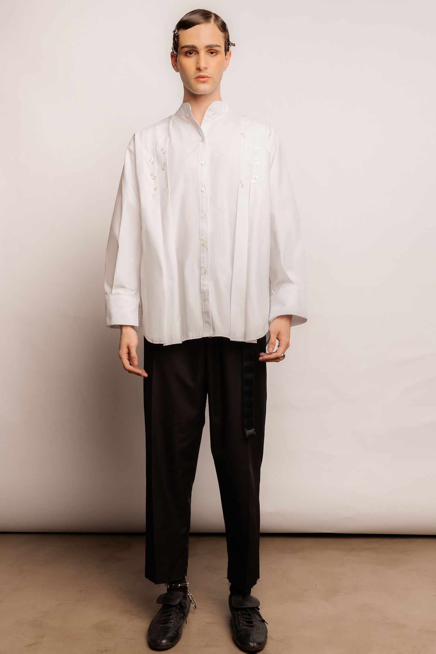 Model in an oversized white pleated button shirt with a mandarin collar, black belted trousers and black leather shoes.