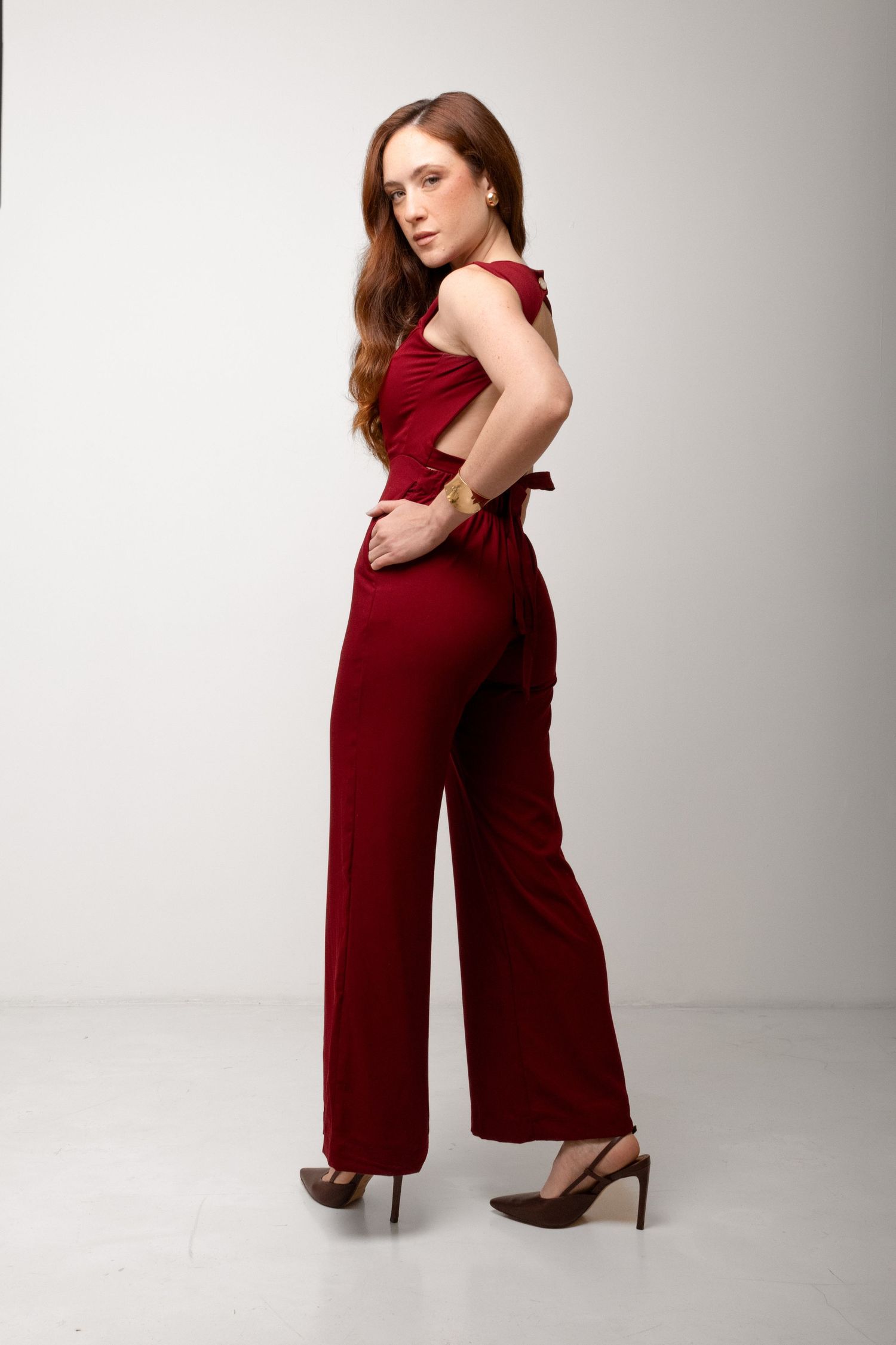 Woman in a burgundy sleeveless jumpsuit with open back detail and wide legs poses in profile wearing dark pointed heels.