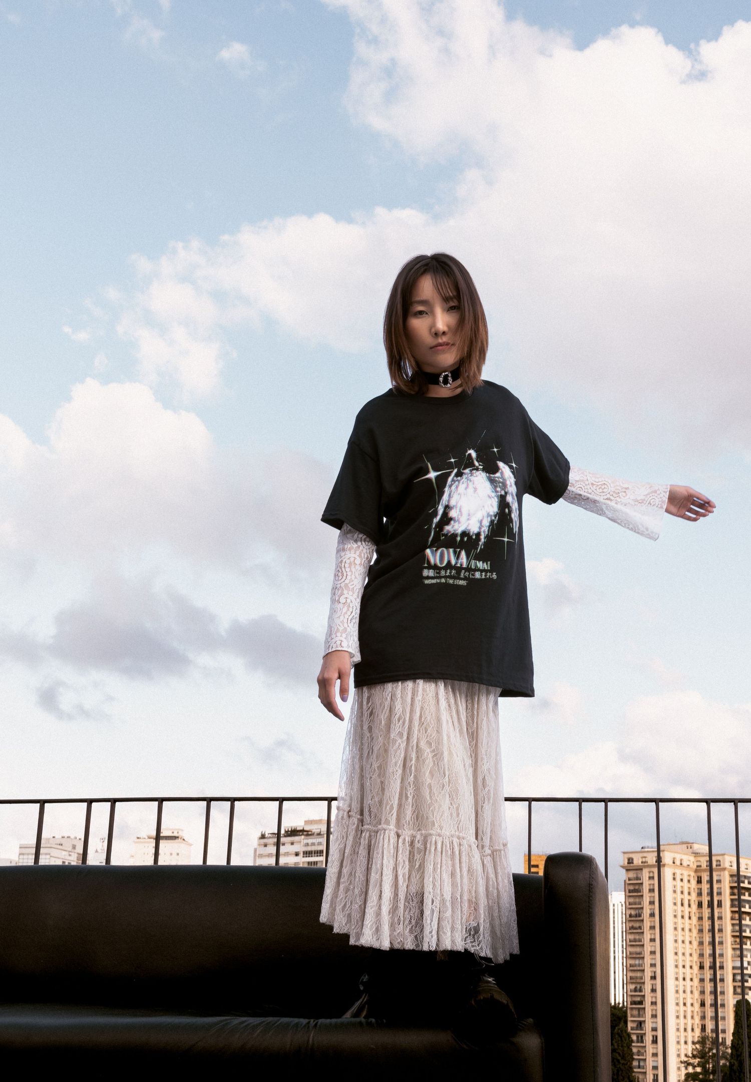 A woman in an oversized black graphic tee and long white lace skirt stands on a rooftop sofa against a cloudy blue sky.