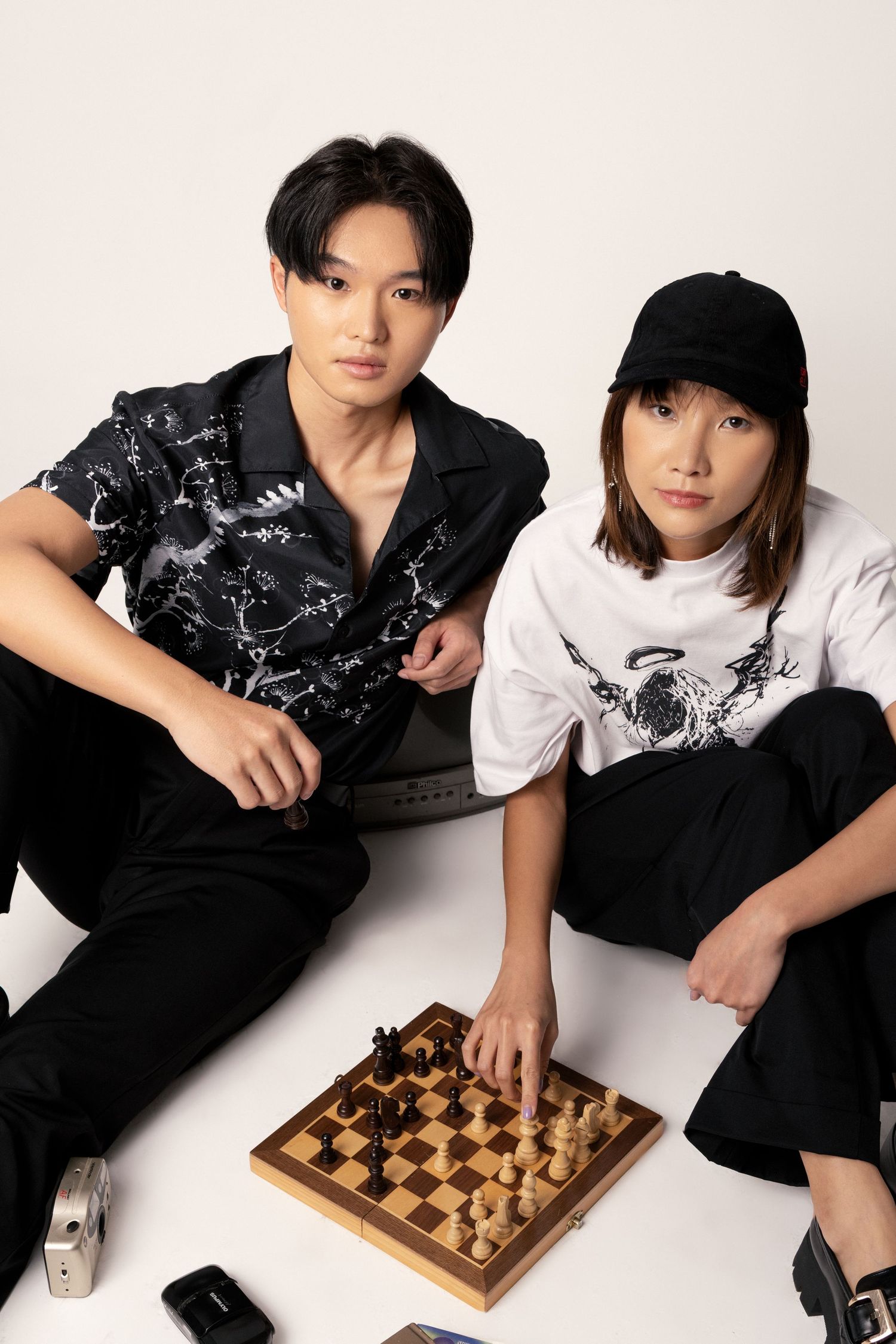 Two young models in black and white streetwear sit on a white floor playing chess on a wooden board.