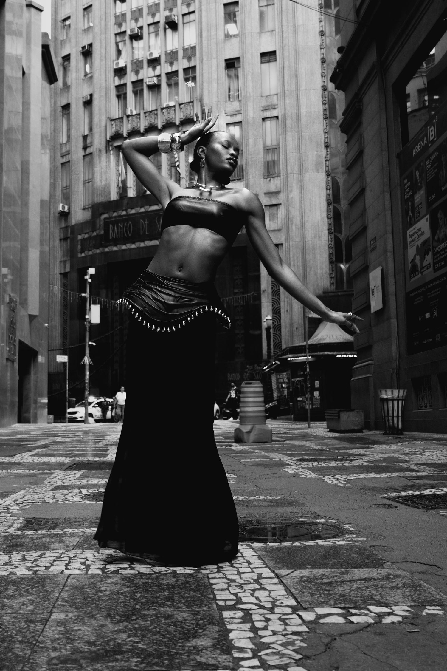 Woman in a black bandeau top and maxi skirt strikes a dance-like pose with raised arm on a mosaic-paved street between tall buildings.