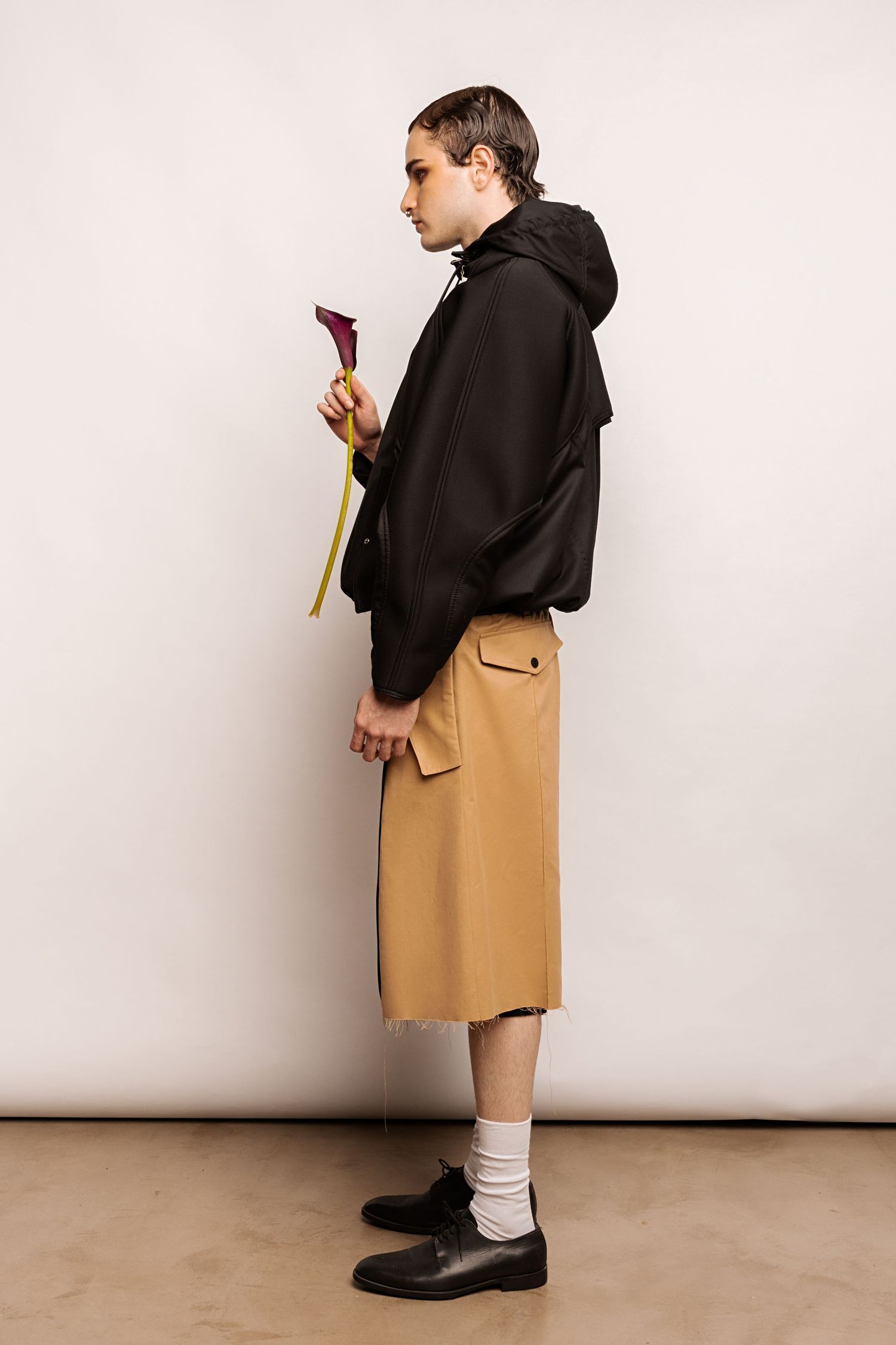 Side profile of a model in a black hooded jacket and tan shorts with a flap pocket, holding a dark calla lily.