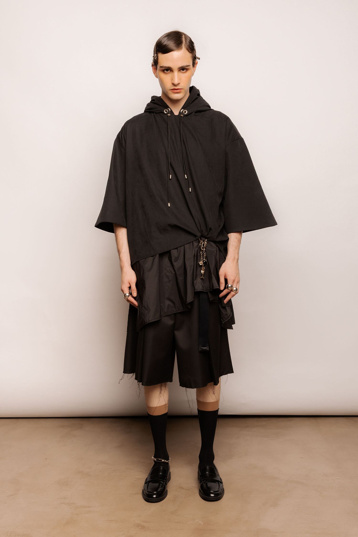 Model in a black oversized hooded cape top layered over black shorts and an asymmetric skirt, with black socks and glossy loafers.