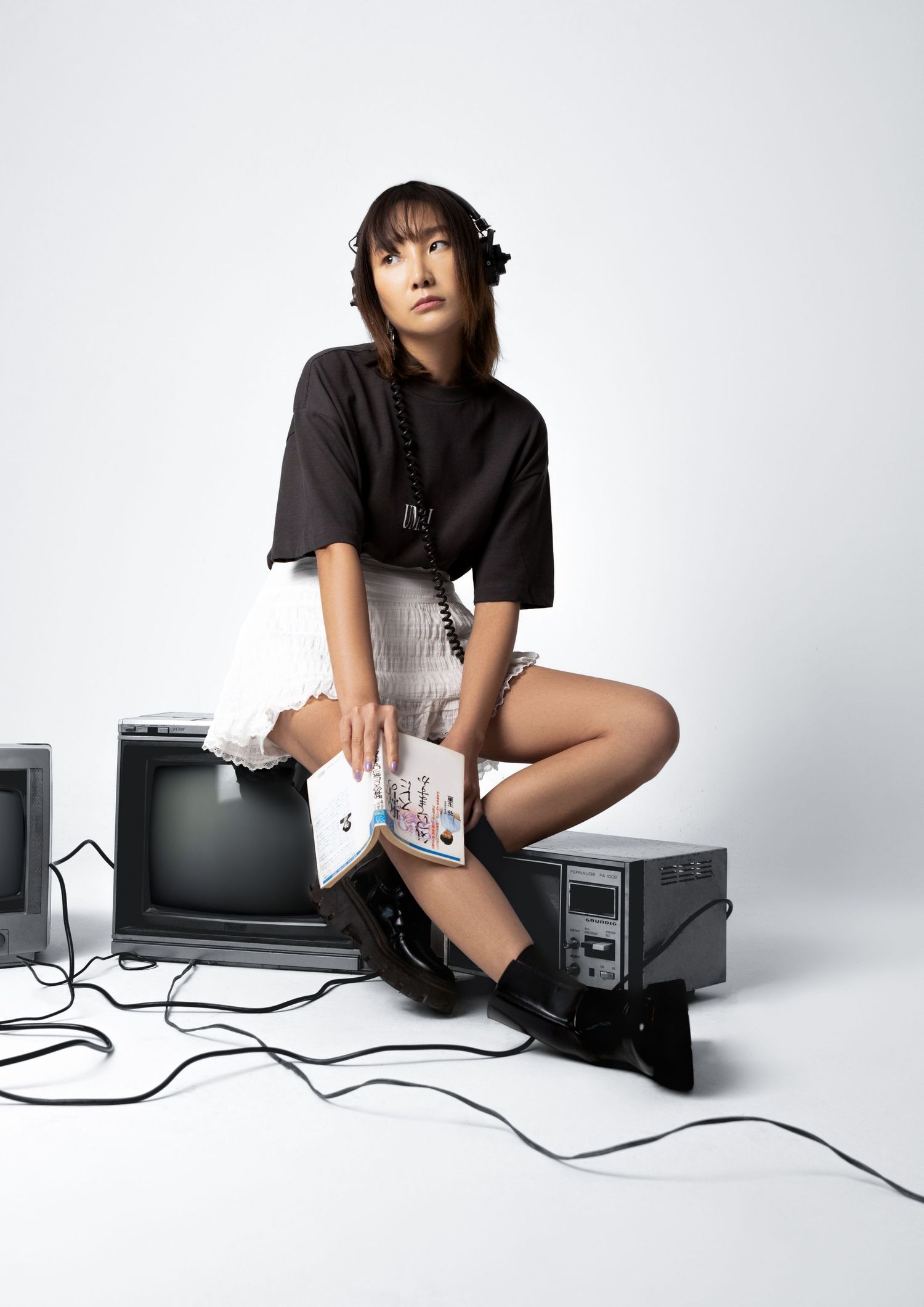 A woman in a black tee, white ruffled skirt and boots sits on retro televisions reading a colorful book.