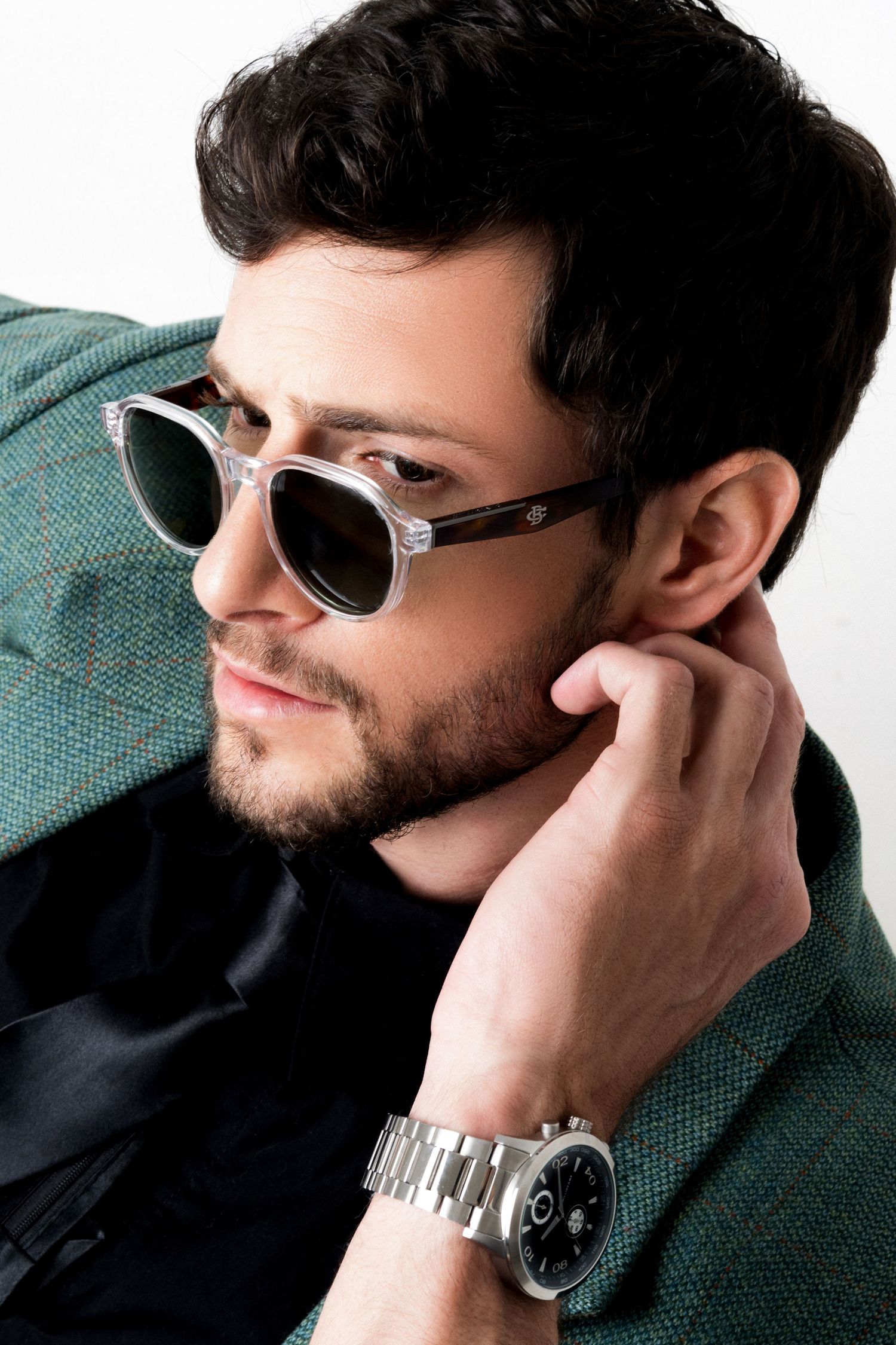 Bearded man in clear-framed sunglasses and a green tweed jacket rests his hand behind his neck, wearing a silver chronograph watch.