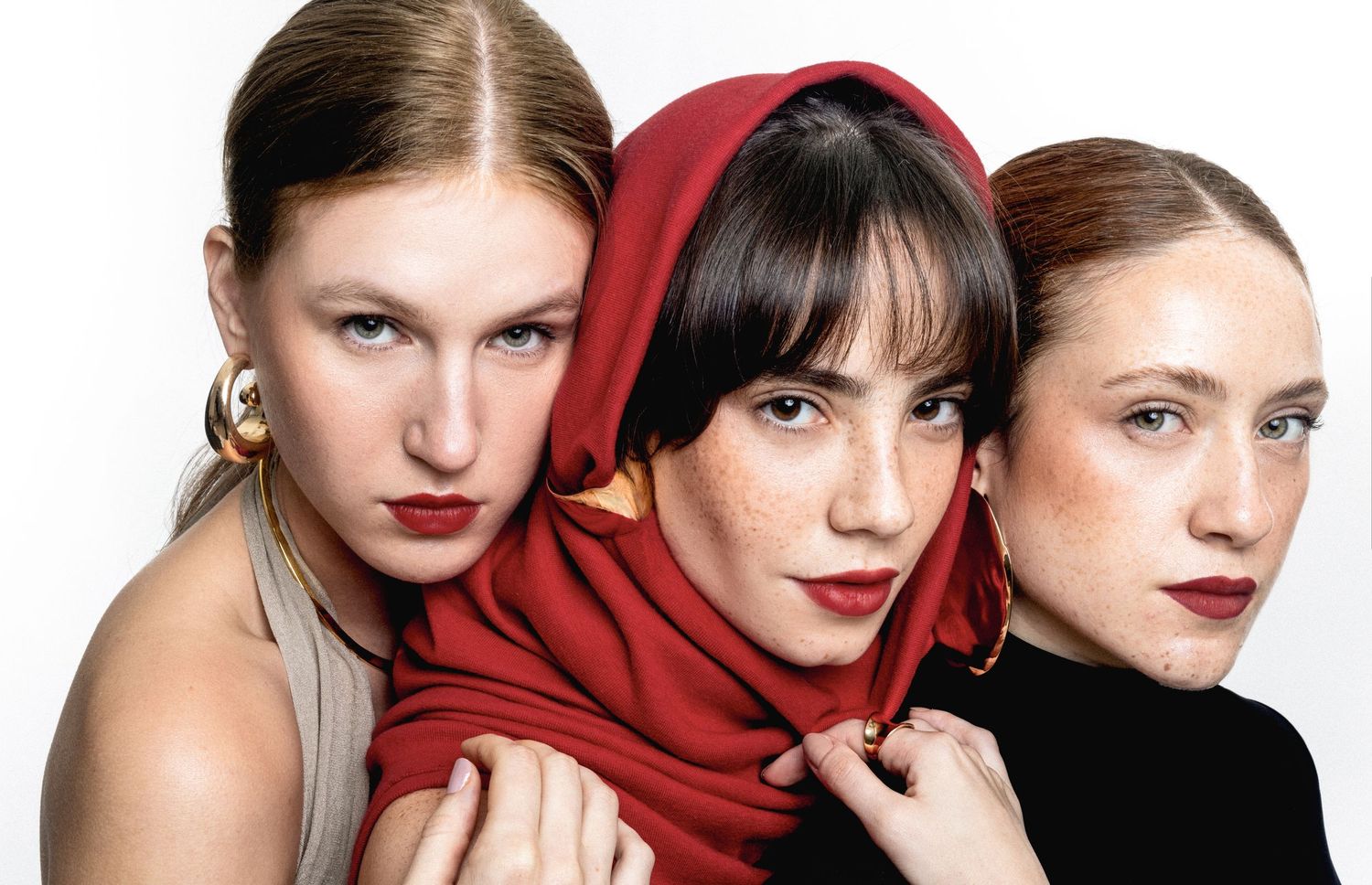 Three women with red lipstick pose closely together, one wearing a red headscarf, in a bold beauty editorial portrait.