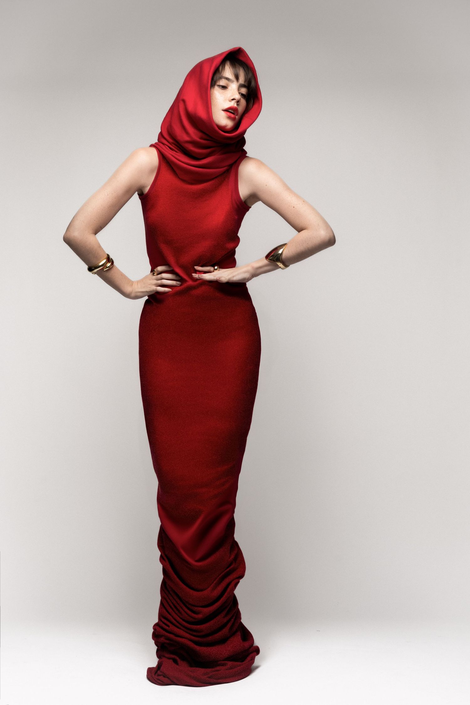 Full-length studio fashion shot of a woman in a floor-length red hooded gown with gold bracelets, hands on hips.
