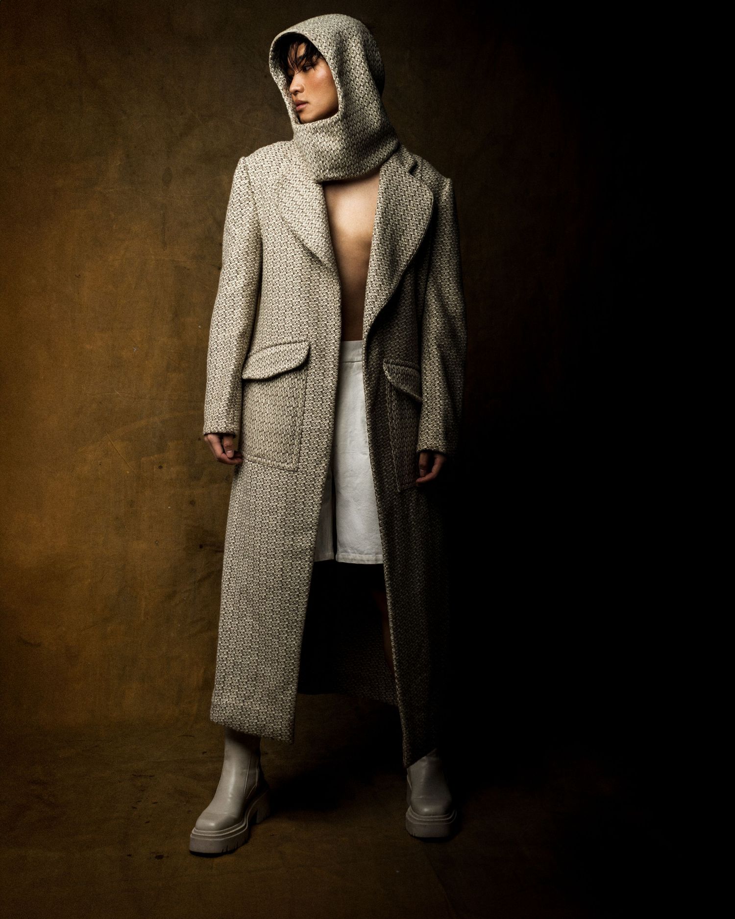Full-length fashion shot of a model in a long hooded tweed coat, white shorts and chunky cream boots against a textured brown backdrop.