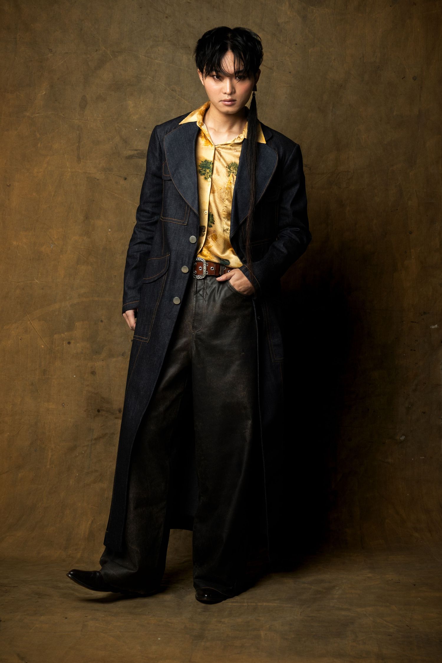 Model in a long dark denim coat, yellow floral shirt and loose leather trousers stands with a hand in pocket against a brown backdrop.