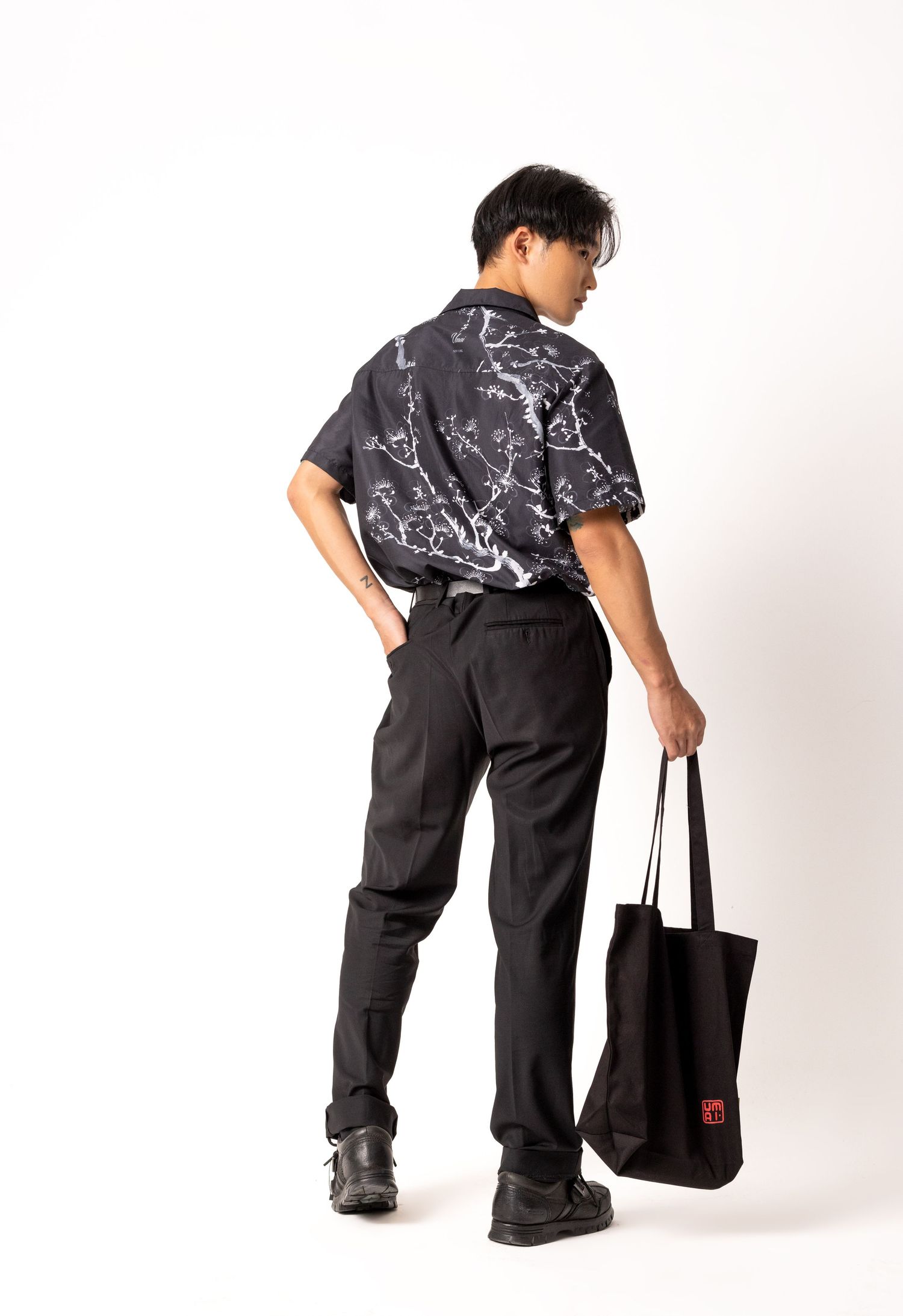 Man seen from behind wearing a black floral-print short-sleeve shirt and black trousers, holding a black canvas tote bag.