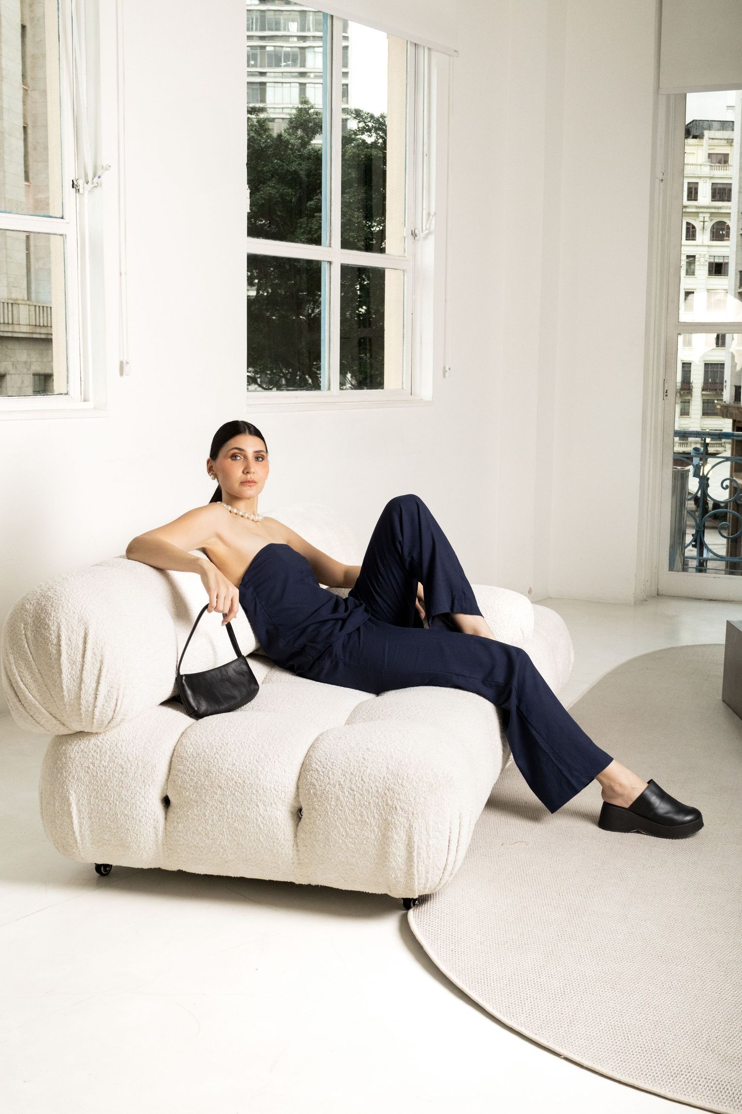 Model in a navy strapless jumpsuit and black loafers lounges on a white bouclé sofa holding a small black shoulder bag.