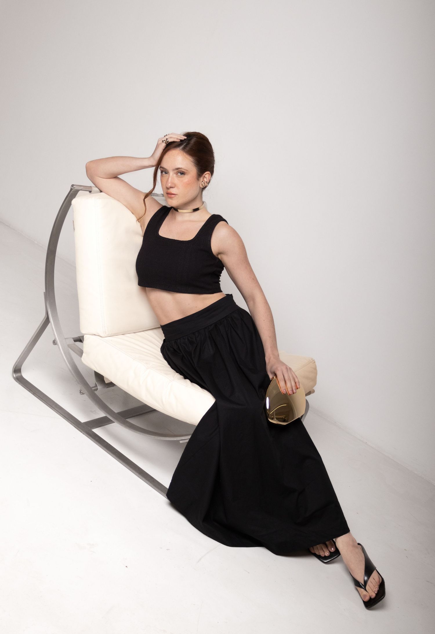 Model sits on a modern cream chair with one hand in her hair, dressed in a black crop top and billowing black maxi skirt.