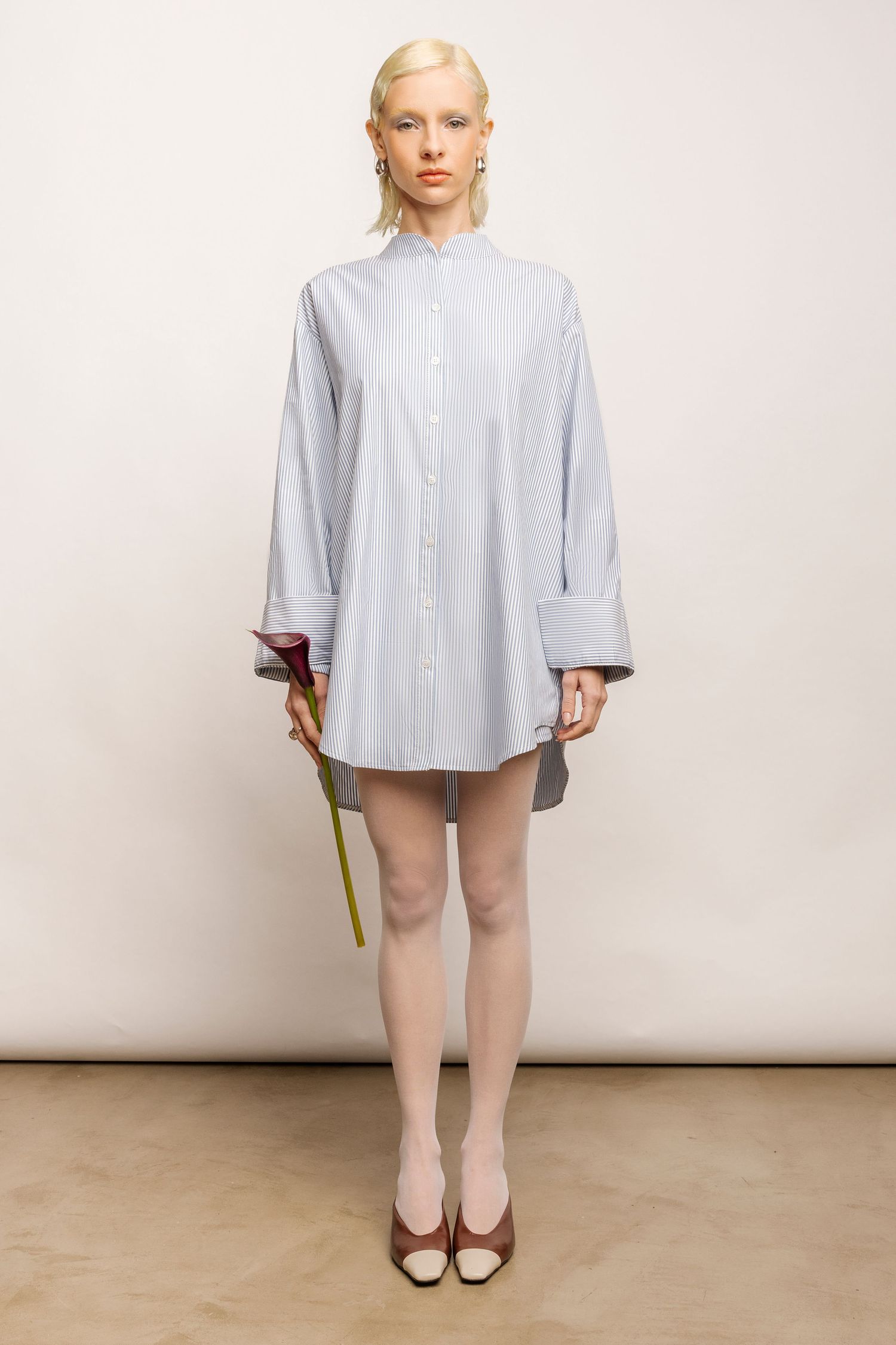 Model in an oversized blue striped button-up shirt dress, sheer tights and two-tone heels holds a calla lily by her side.